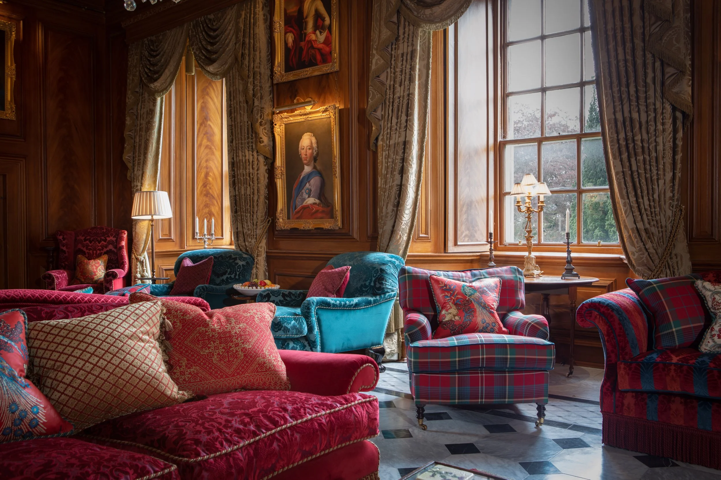 A luxurious living room with ornate wooden walls, large windows with draped curtains, and colorful velvet armchairs and sofas. Walls are decorated with framed classical portraits, and a chandelier with lamps provides warm lighting.