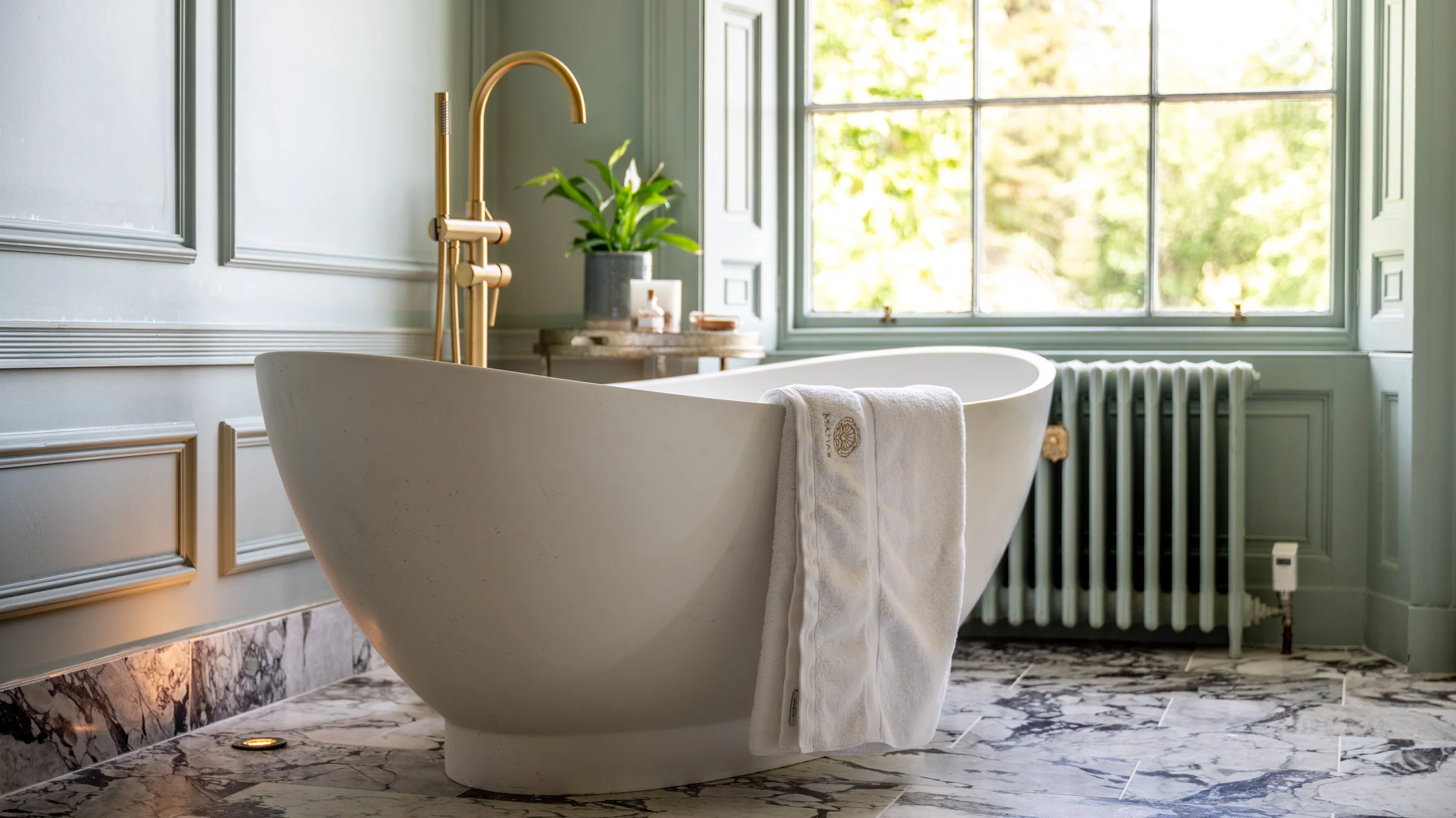 A luxurious bathroom featuring a standalone white tub with a white towel draped over its edge, set on a marble floor. A large window with greenish-blue trim allows natural light to fill the room, and a radiator is visible beneath the window. A small 