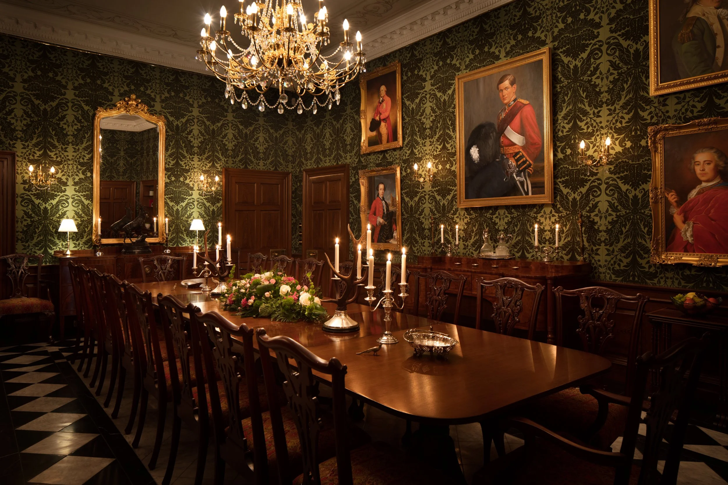 Elegant dining room with a large chandelier hanging from the ornate ceiling, decorated with framed portraits on the dark patterned wallpaper, and a long wooden table set with candles and a floral centerpiece.