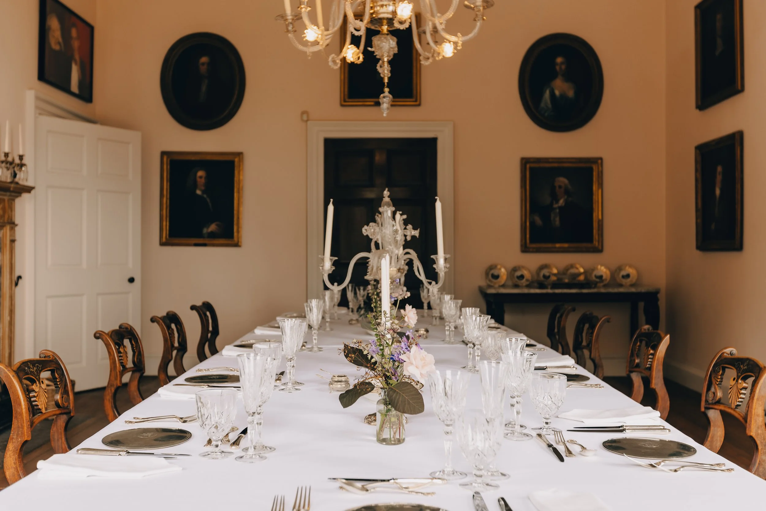 A long dining table set for a formal meal with a white tablecloth, glassware, silverware, and a floral centerpiece. The room has a chandelier, framed portraits, and elegant wooden chairs.