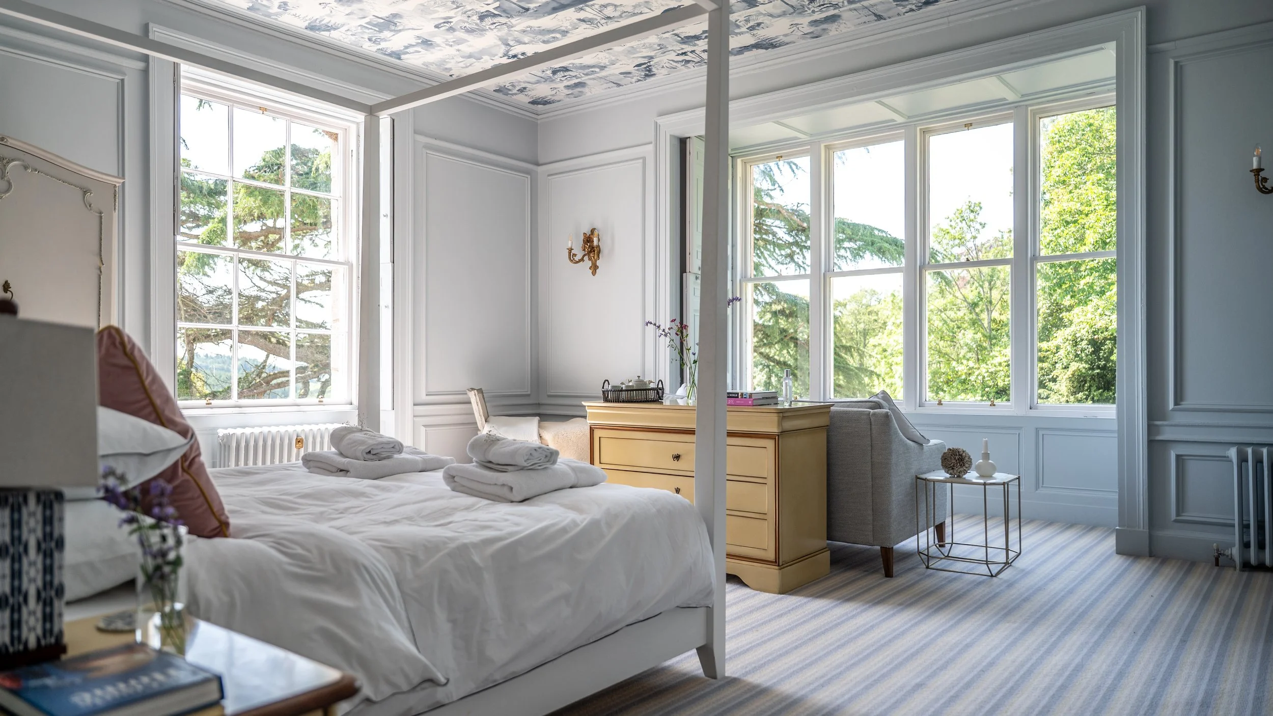 A bright and elegant bedroom with large windows showing green trees outside. The bed is made with white linens and has folded towels on top. There is a yellow dresser, a beige armchair, and a small side table with decorative items. The room has light