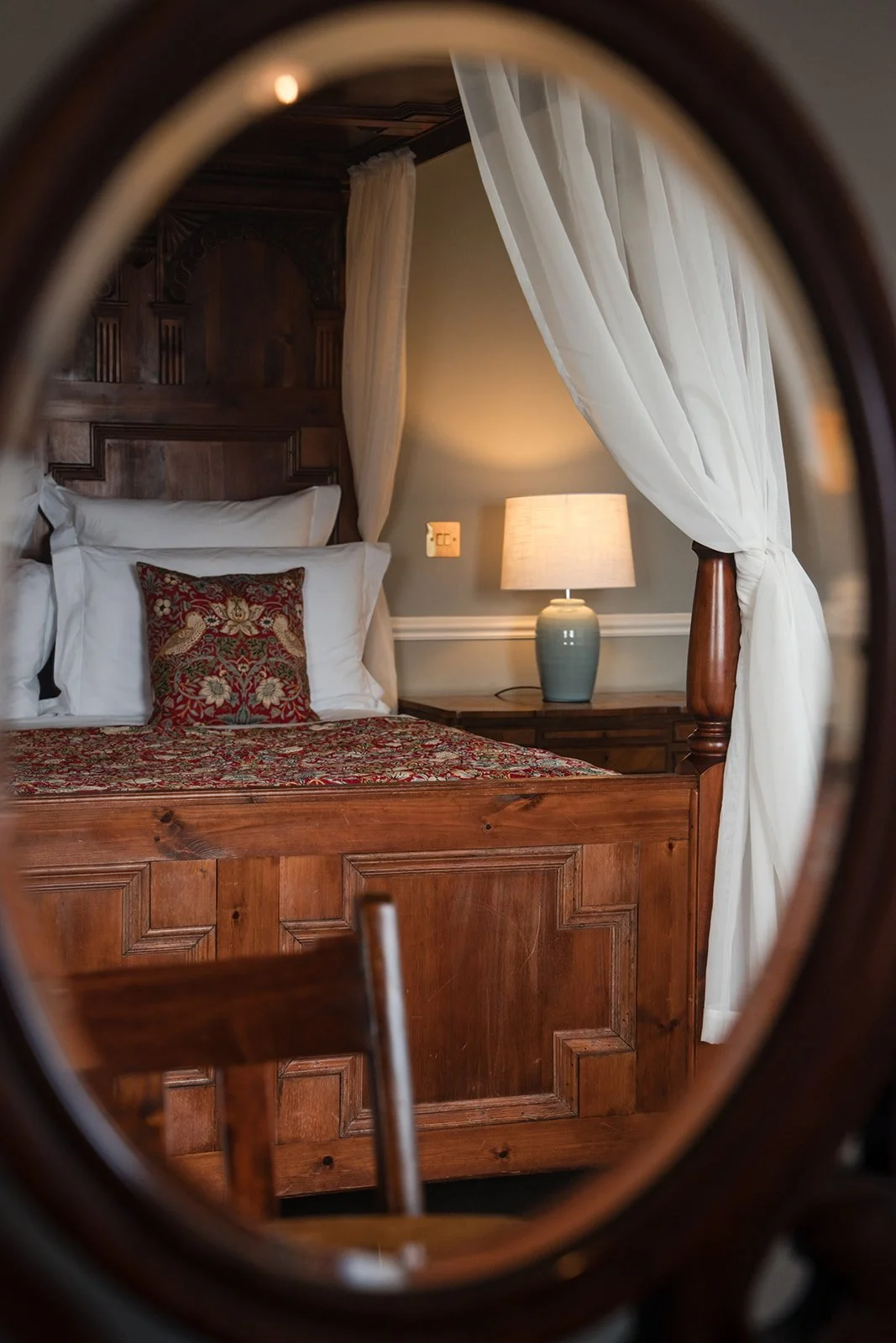 A view of a bedroom seen through an oval mirror. The bed has white pillows, a decorative pillow with a floral pattern, a wooden headboard, and a matching wooden bed frame. A bedside table with a blue lamp and a wall with a beige color and white trim 