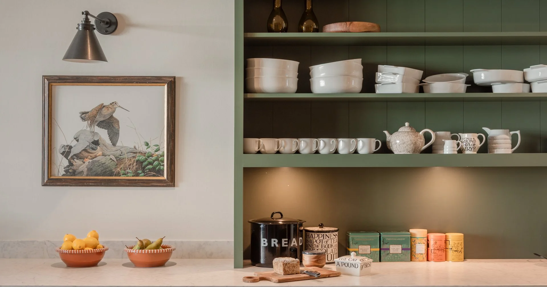 Part of a kitchen with open green shelves filled with white bowls, cream-colored cups, and teapots. On the countertop, there are two bowls with lemons and pears, a black bread container, a tea canister, and colorful tea tins. There's a framed picture