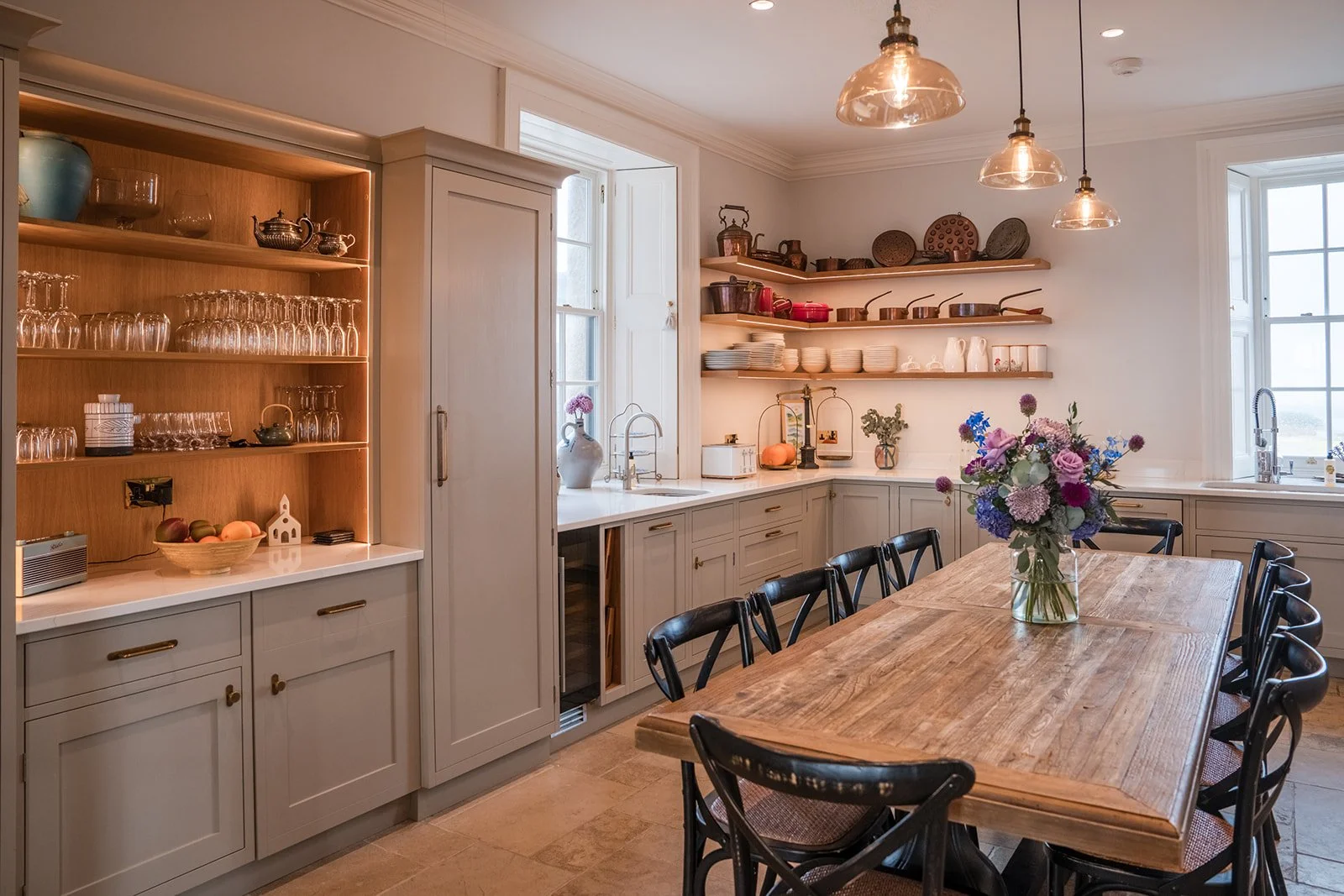 Modern kitchen with wooden table, black chairs, open shelves with dishes, vases, flowers, and large windows.