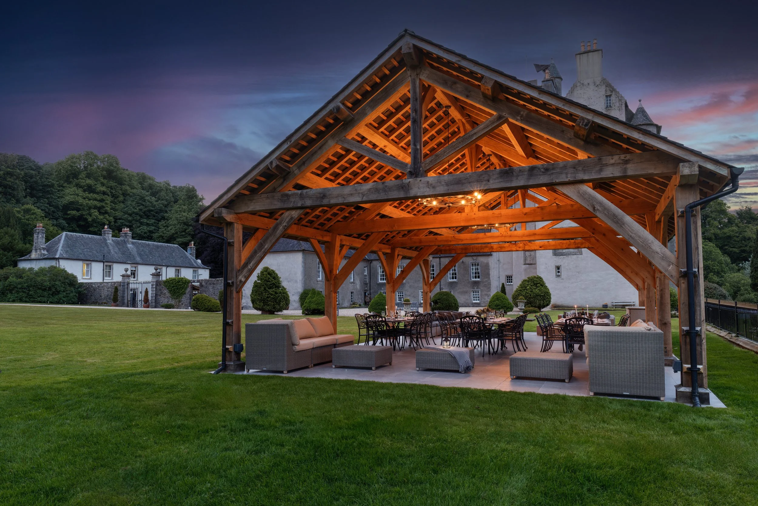 An outdoor patio area with a wooden pergola and seating, overlooking a historic castle-like building at dusk or early evening.