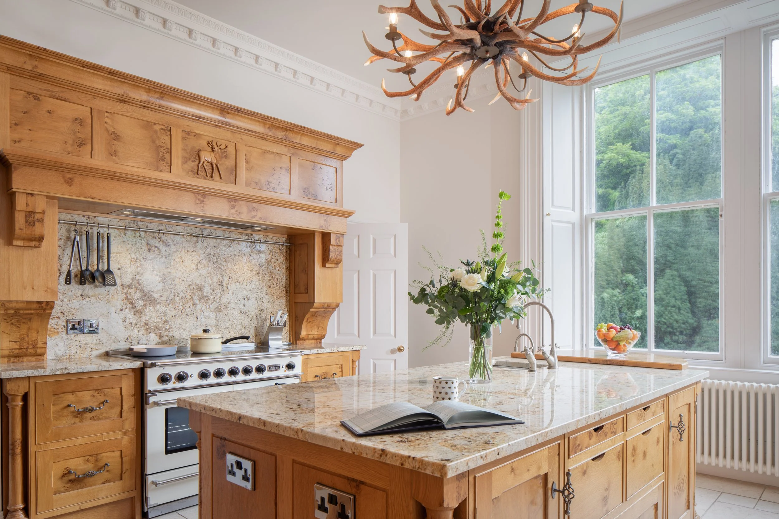A kitchen with wooden cabinets, granite countertops, a stove, and a large window with a view of trees. There is a vase of flowers and a bowl of fruit on the counter, and a decorative antler chandelier hanging from the ceiling.