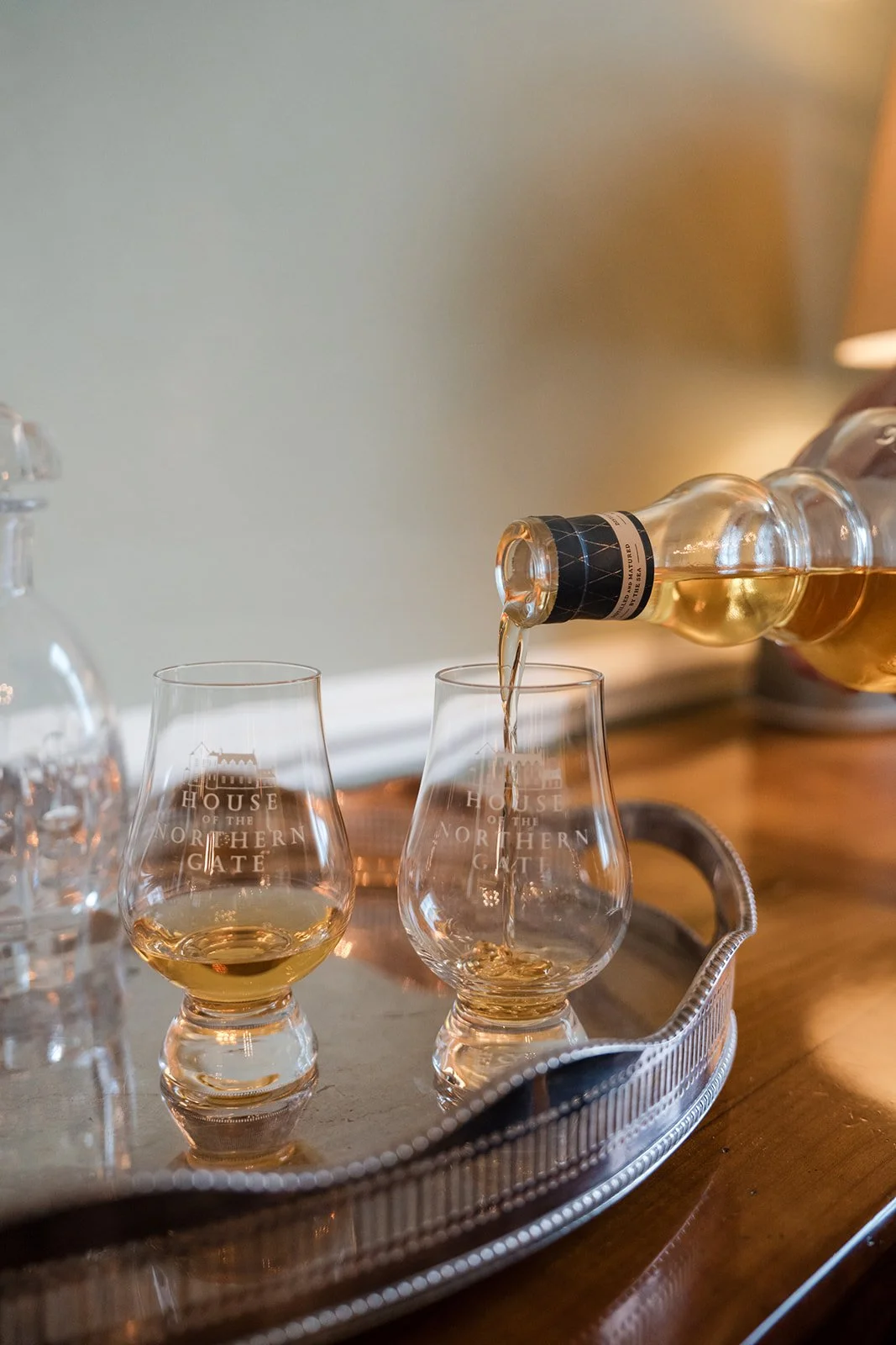 A bottle of whiskey pouring into a glass with the logo 'House of the Northern Gate' on it, on a silver tray with other glasses, in a cozy setting.