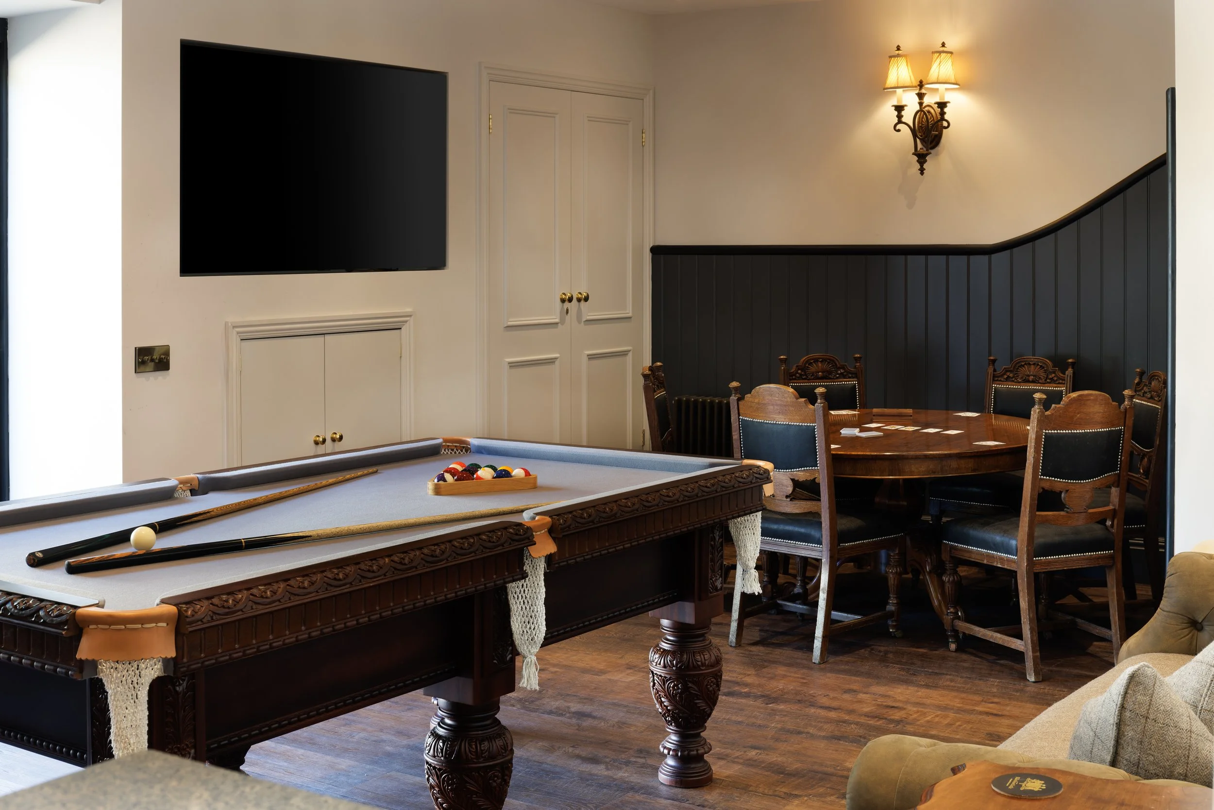 A room with a pool table and a drinking table, dark wood furniture, and a wall-mounted TV. There are chairs around the table, a dark blue and wood wainscot on the wall, an elegant wall sconce with two lights, and a closed white cabinet door.