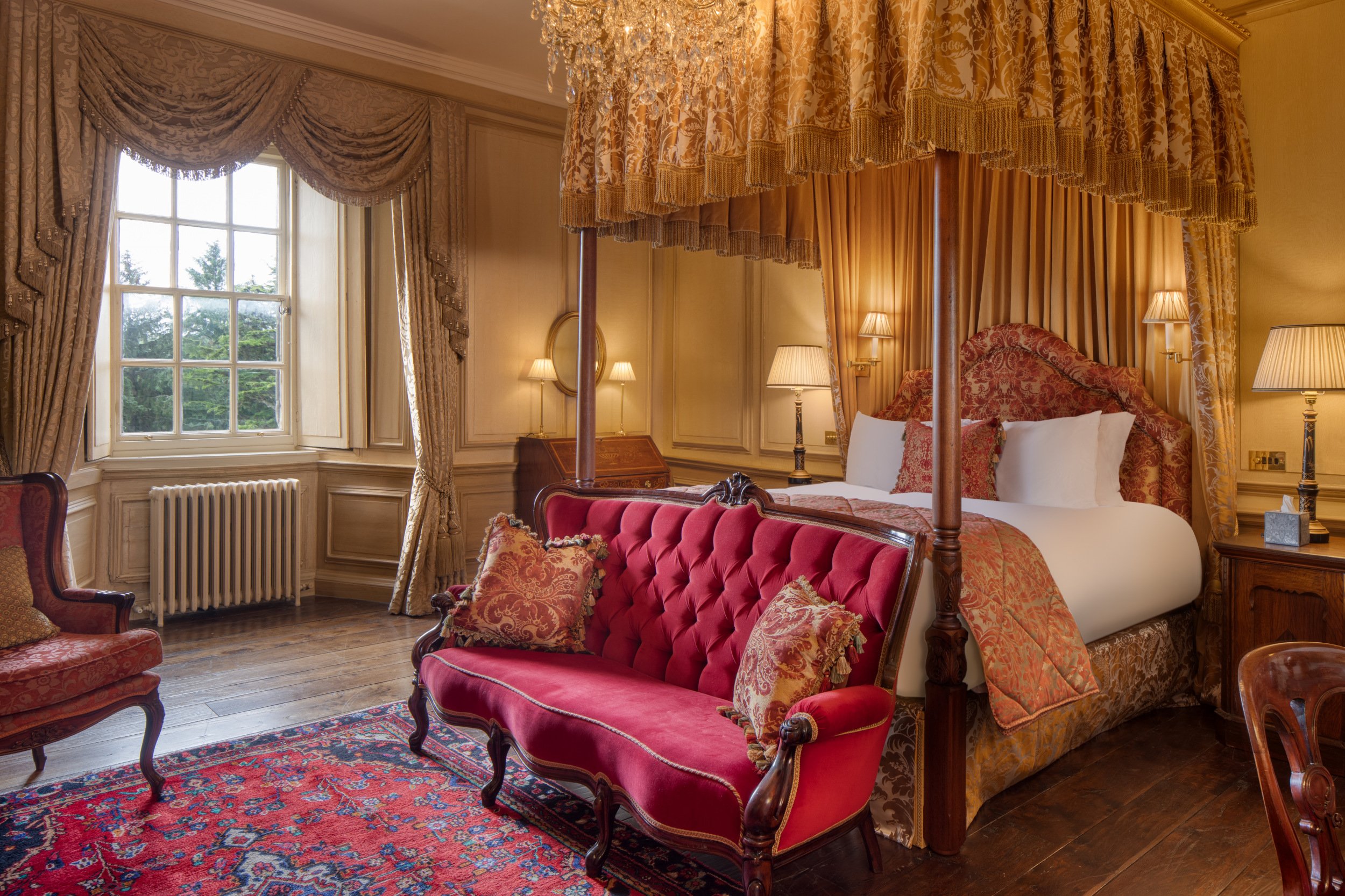 A luxurious bedroom with a four-poster bed with gold and patterned drapes, a red velvet tufted loveseat, antique wooden furniture, a window with ornate curtains, and warm lighting from table lamps and a chandelier.