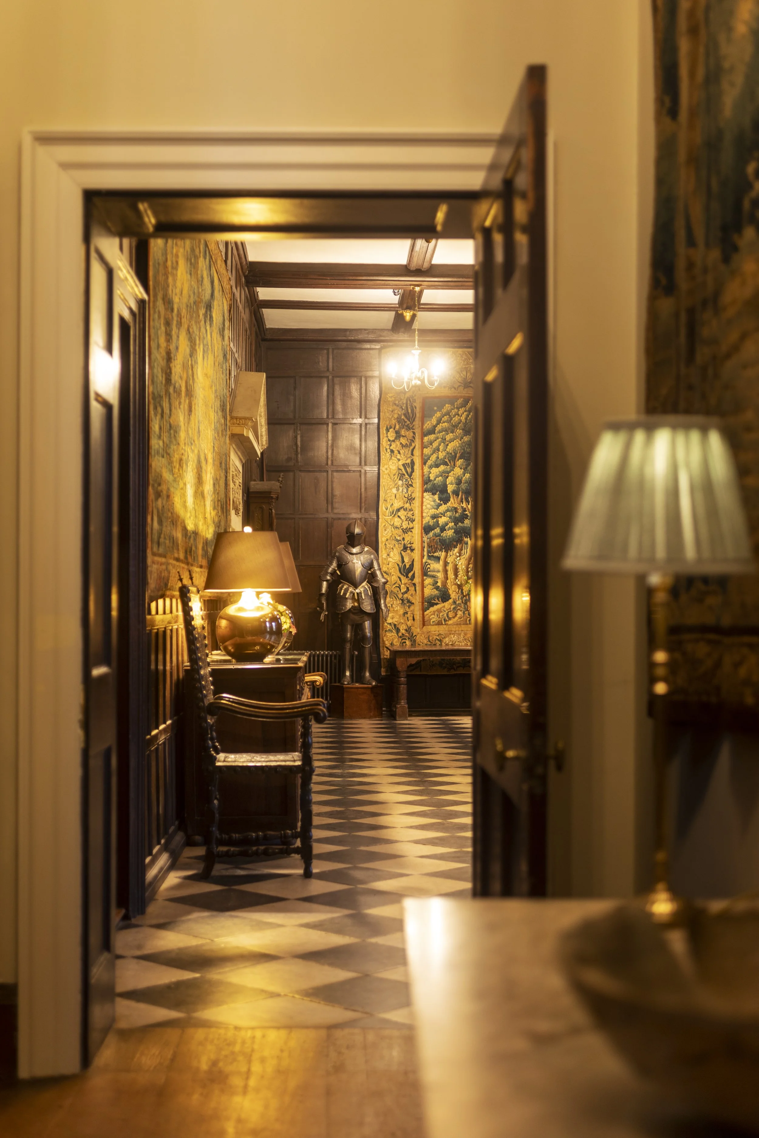 A hallway with checkerboard black and white flooring, wooden paneled walls, and antique furniture. A suit of armor stands in the background, and a chandelier hangs from the ceiling. Warm lighting creates a cozy, vintage atmosphere.