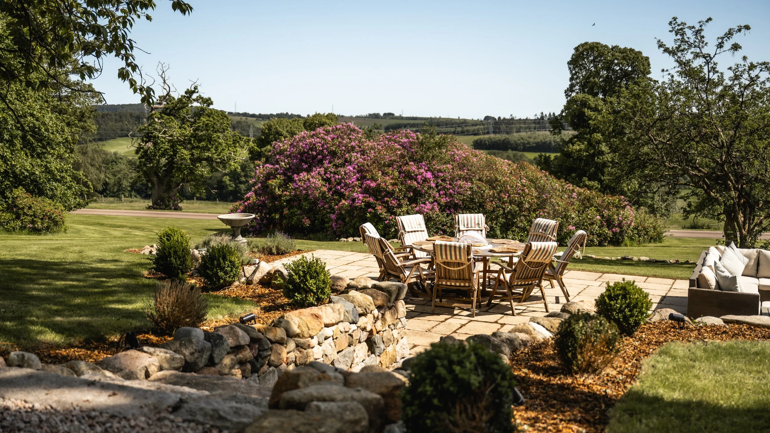 A backyard patio with outdoor dining table and chairs, surrounded by green grass, bushes, and trees, overlooking a hilly countryside with pink flowering bushes and a clear blue sky.