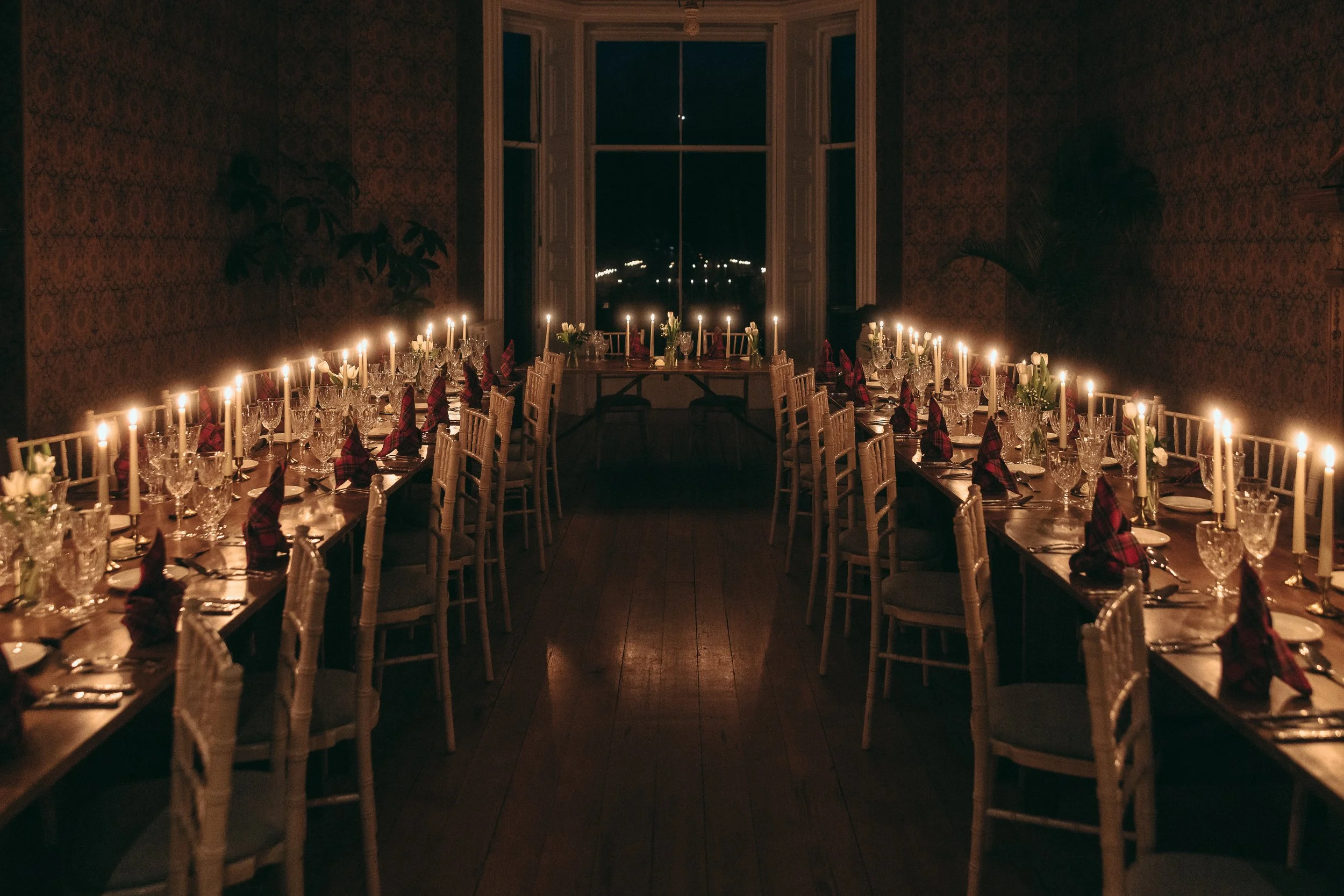 A dimly lit dining room with two long tables set for a formal dinner, decorated with candles, napkins, and glasses, with large windows showing a dark night outside.