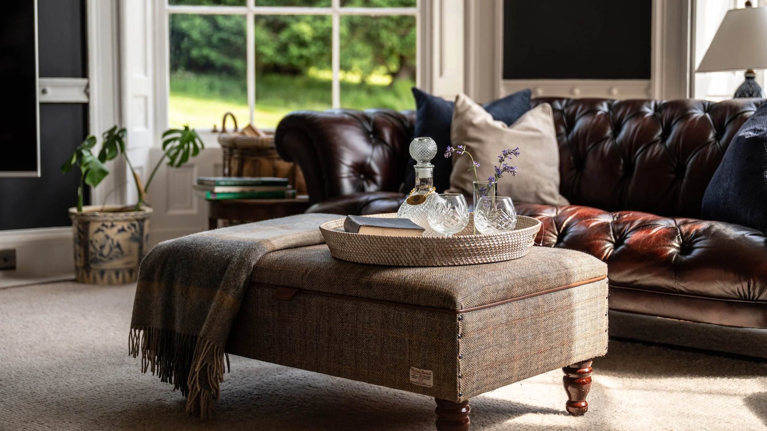 Living room with a large window showing green trees outside, a leather sofa with pillows, a beige ottoman with a tray holding glassware and flowers, and a potted plant, decorated in neutral and dark tones.