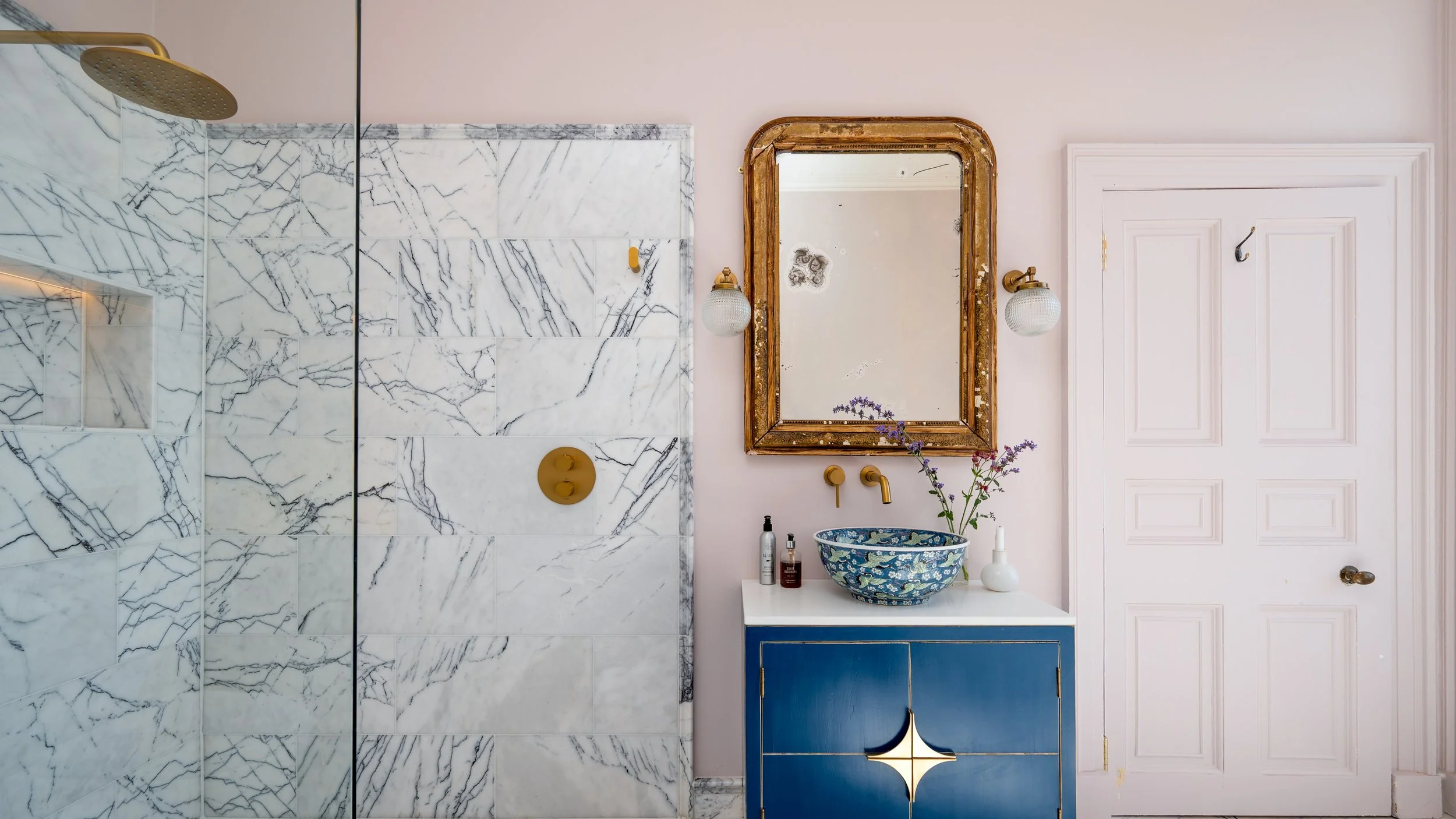 A bathroom with a marble shower, an ornate gold-trimmed mirror, wall-mounted lights, a vanity with a blue cabinet, a patterned basin, and various toiletries.