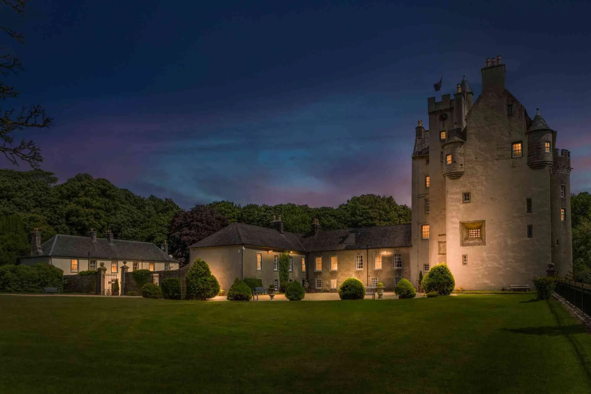 A large castle at dusk with illuminated windows, surrounded by trees and smaller buildings, under a darkening sky.