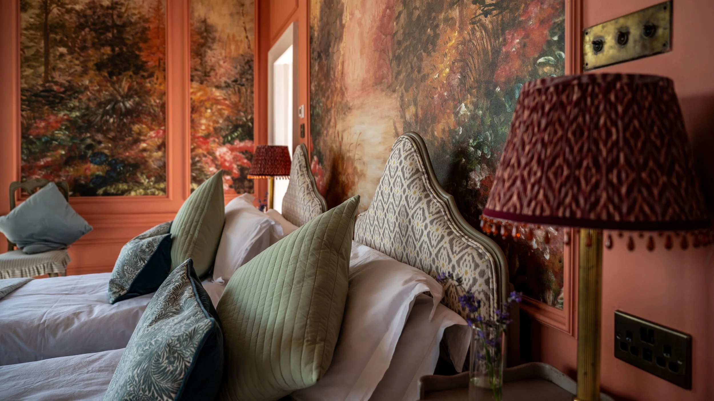 A close-up of a bedroom with a bed, pillows, and a vibrant mural of an autumn landscape on the wall behind the headboard. Lamps on bedside tables with patterned shades.
