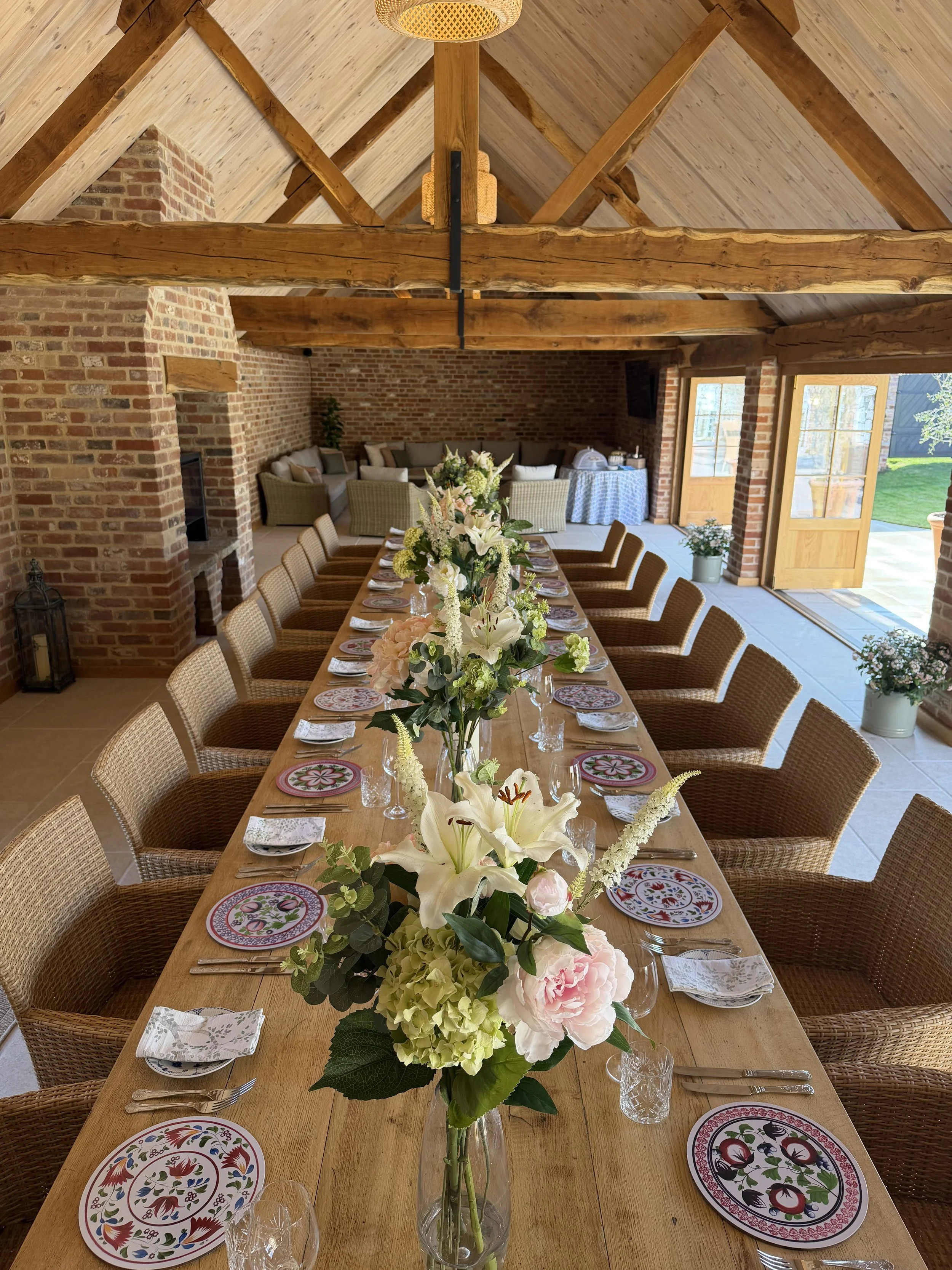 A long wooden dining table set for a meal with floral plates, glasses, cutlery, and a large bouquet of white and pink flowers in a glass vase. The room has exposed brick walls, wooden beams, large windows, and a doorway leading outside to a grassy ar