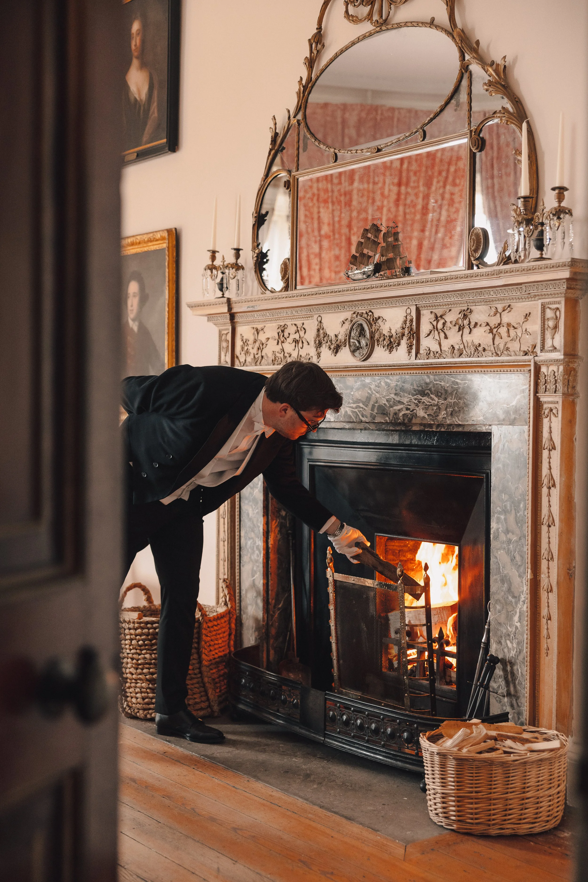 A butler in a suit bending over to adjust a fire in a decorative fireplace with an ornate gold mirror above it. The room has decorated walls with portraits, and there are baskets containing wood nearby.