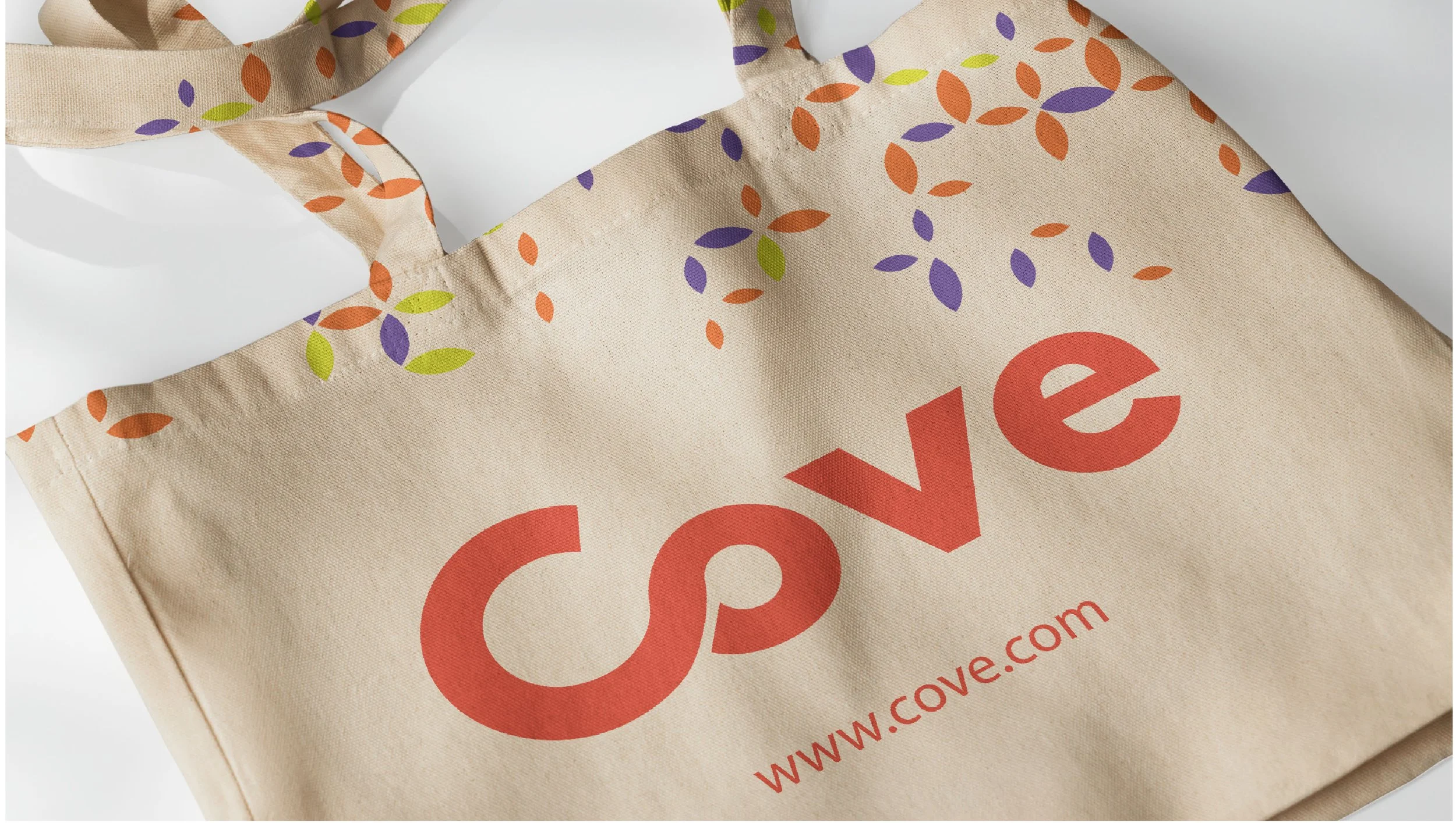 Close-up of a beige reusable tote bag with handles, featuring red text that says 'Cove' and the website 'www.cove.com', and a colorful leaf pattern in green, purple, and orange.