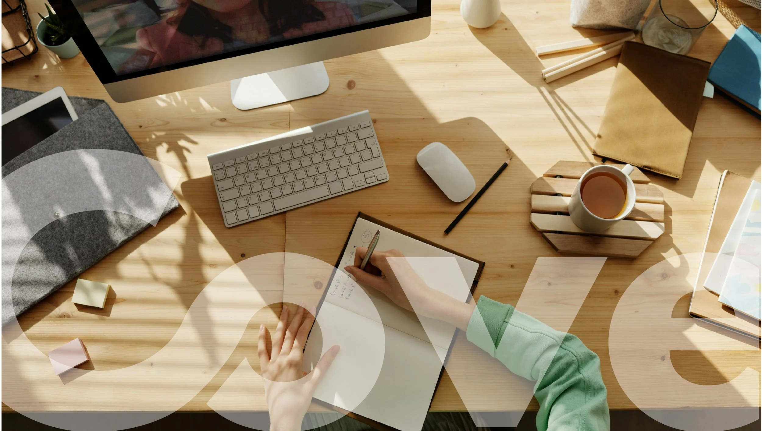A workspace with a wood desk, a computer monitor, keyboard, mouse, a cup of tea or coffee, notebooks, sticky notes, and a person writing in a notebook with sunlight casting shadows.