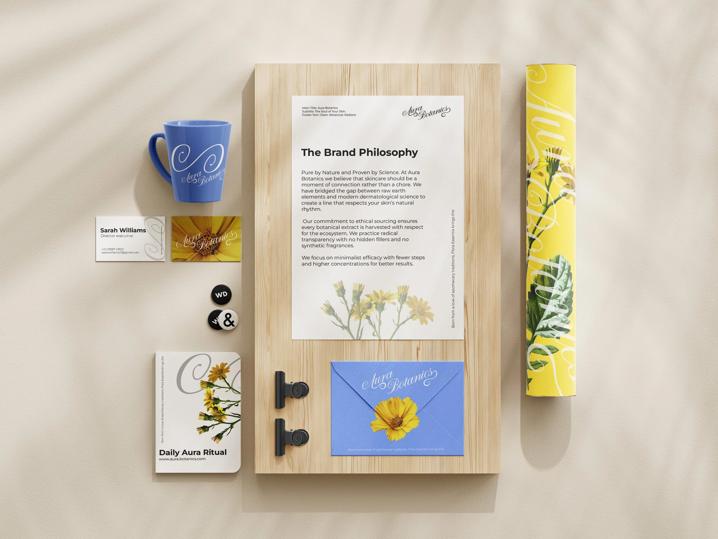 Flat lay of branding and stationery items for Aura Botanics, including a blue mug, business cards, black and white badges, a booklet, a yellow cylindrical container, and a blue envelope, arranged on a light wooden board.