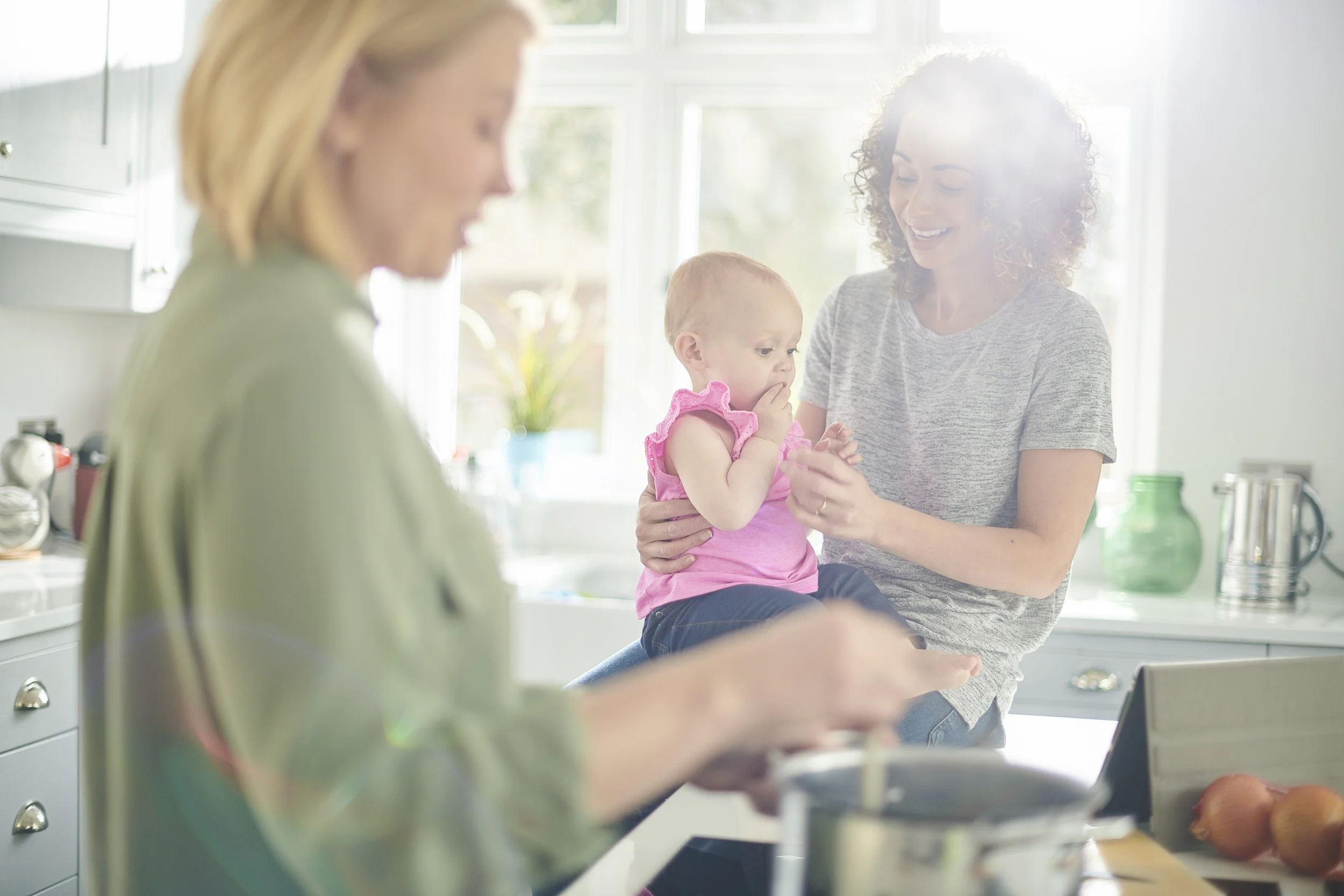 A woman holding a young girl in a kitchen, smiling and engaging in a cooking activity with another woman who is preparing food on the counter.