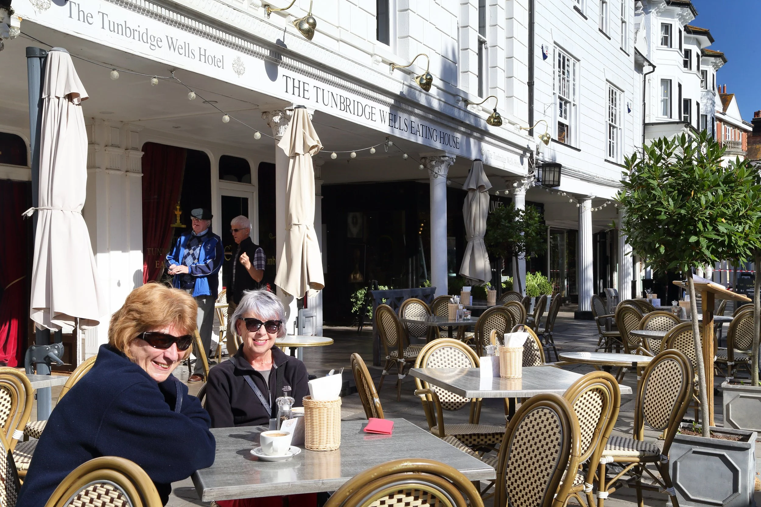 Two women sitting at an outdoor café table, smiling and wearing sunglasses, with a coffee cup on the table, in front of a white building labeled 'The Tunbridge Wells Hotel' with additional signage indicating 'EATING HOUSE'. Other empty tables and cha