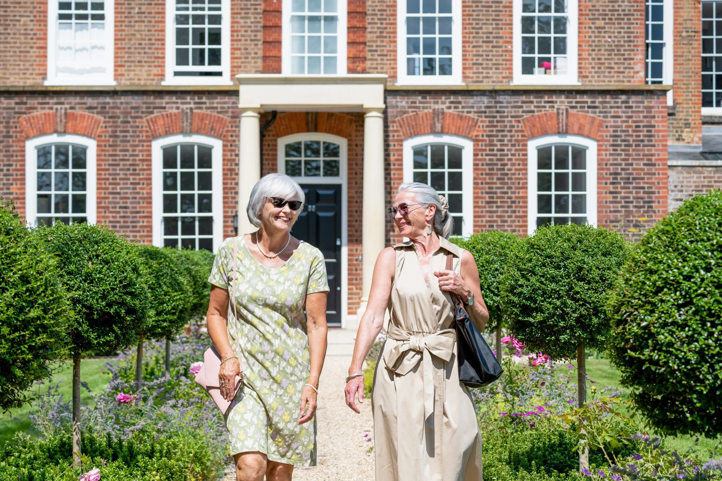 Two elderly women walking and talking in front of a red brick house with white windows and well-maintained garden.