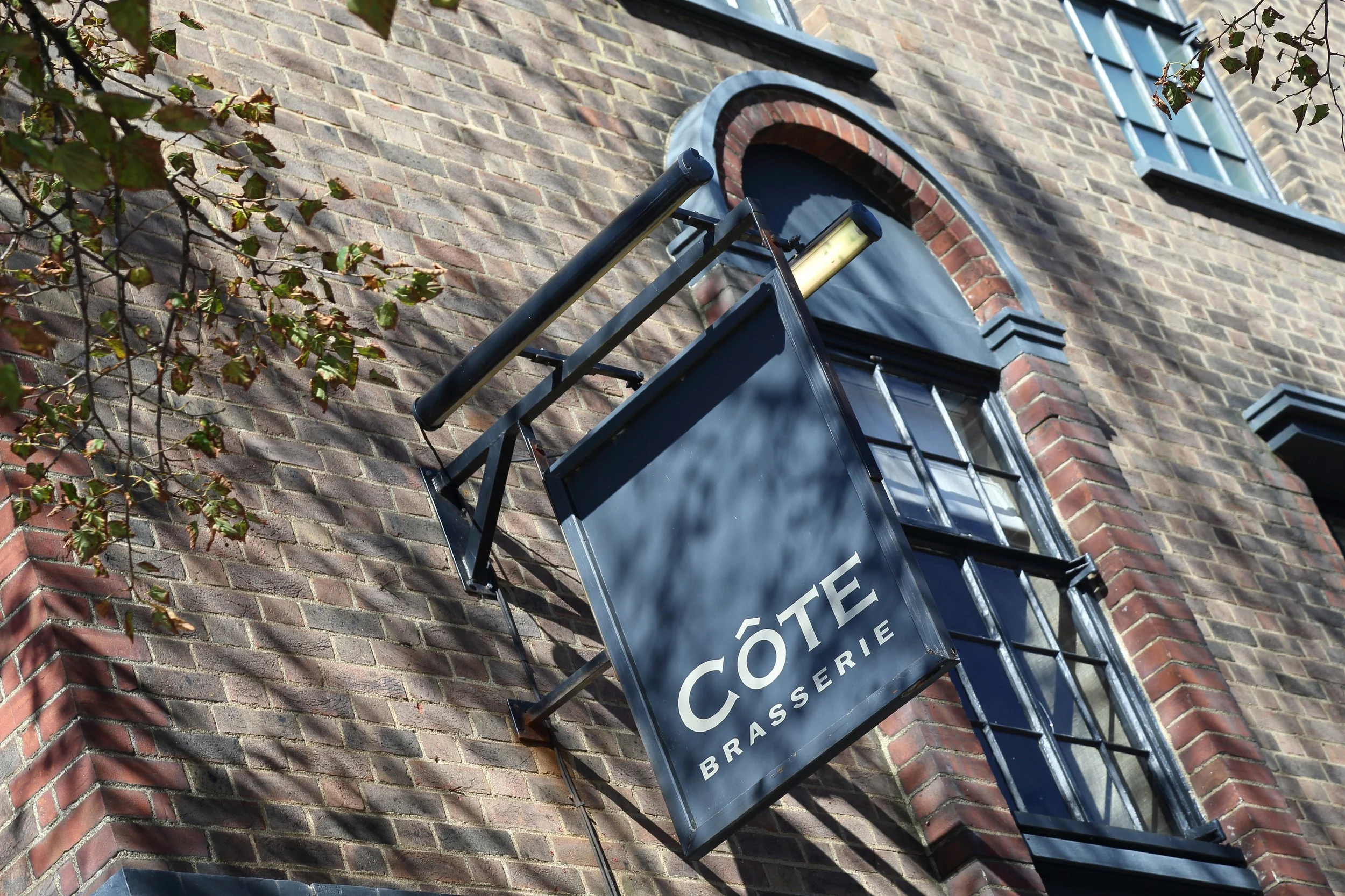 Exterior of a brick building with a hanging sign that reads 'Côte Brasserie' under an arched window.