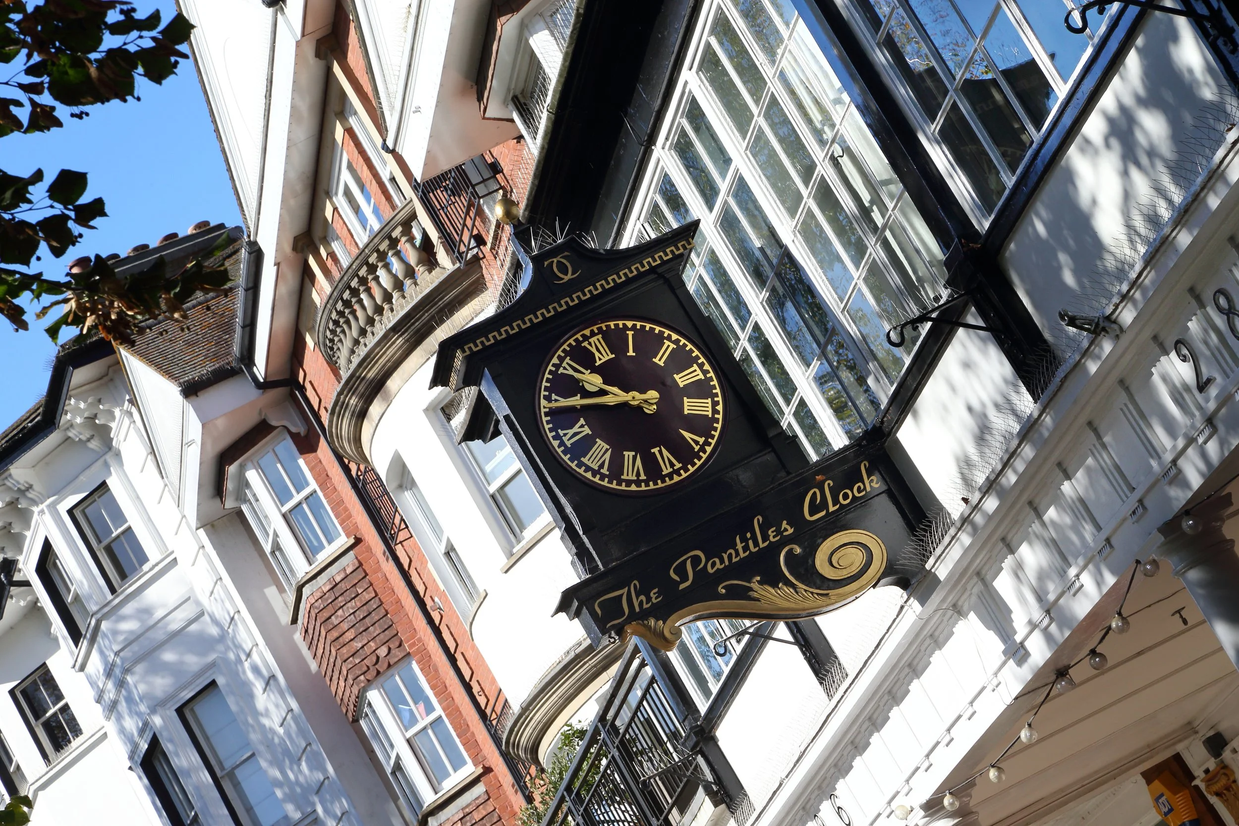 A street view showing a black vintage clock with gold Roman numerals and hands, attached to a building facade with the inscription 'The Pantiles Clock' on a decorative sign below the clock face. The background includes historic buildings with sash wi