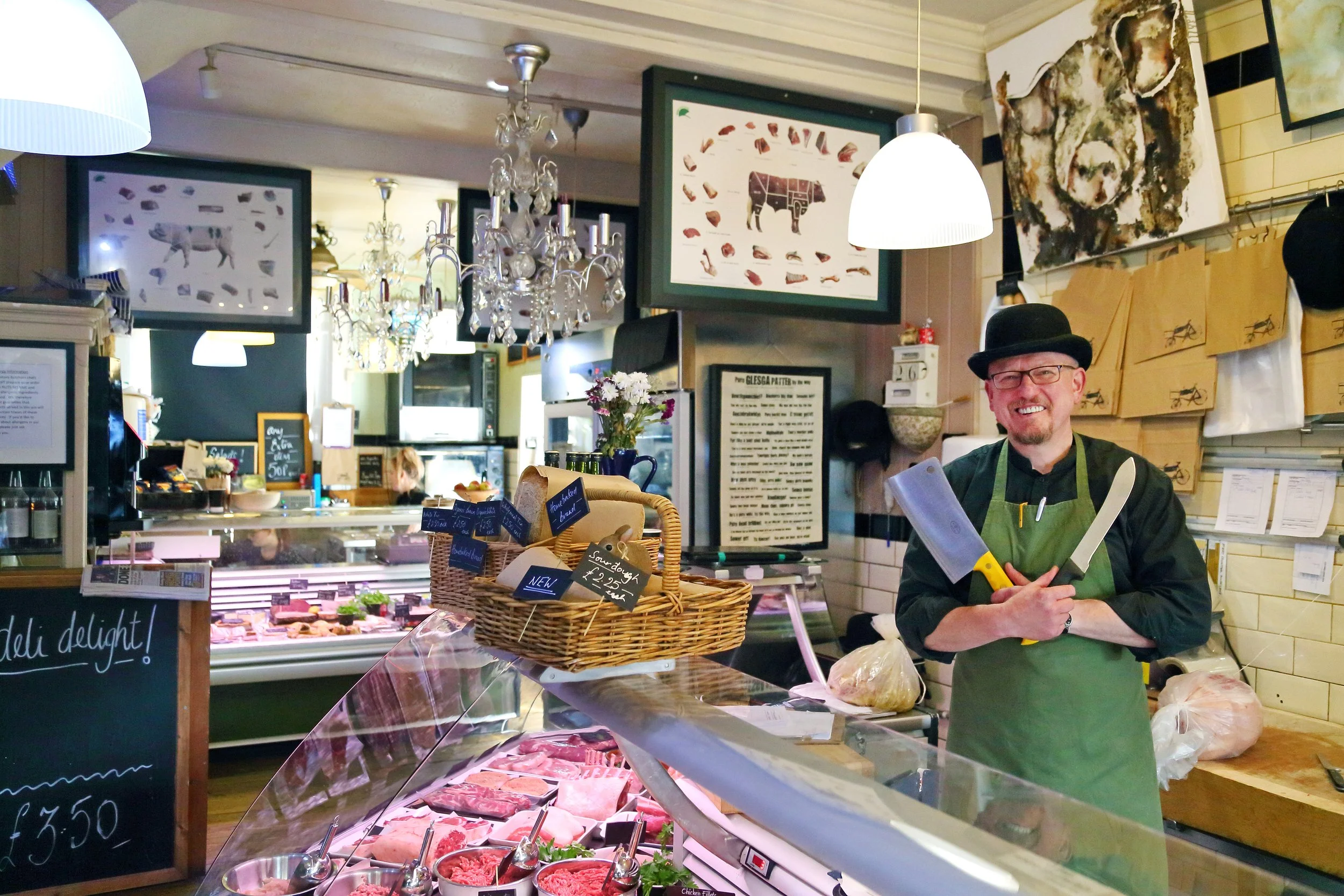 A butcher shop with a smiling butcher wearing a black hat, green apron, and glasses, holding a large knife and a meat cleaver. The shop has a display case filled with various cuts of meat, a basket with small hang tags, and framed pictures of animals
