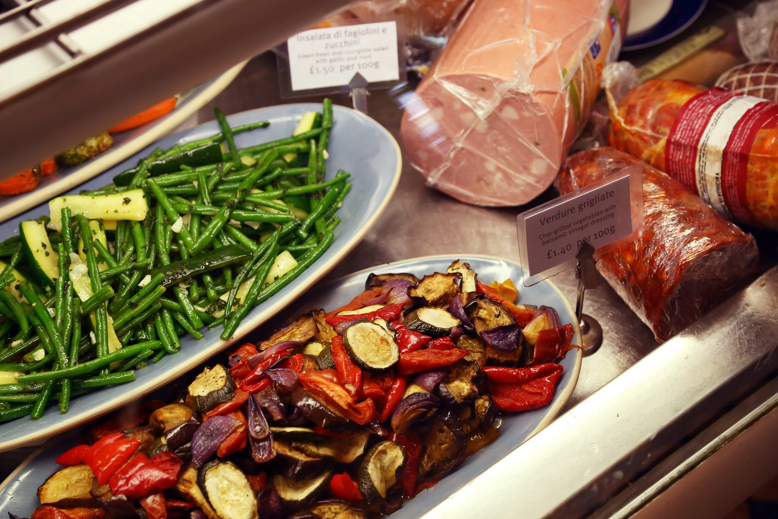 Fresh green beans and chopped roasted vegetables including eggplant and red peppers on display at a market.