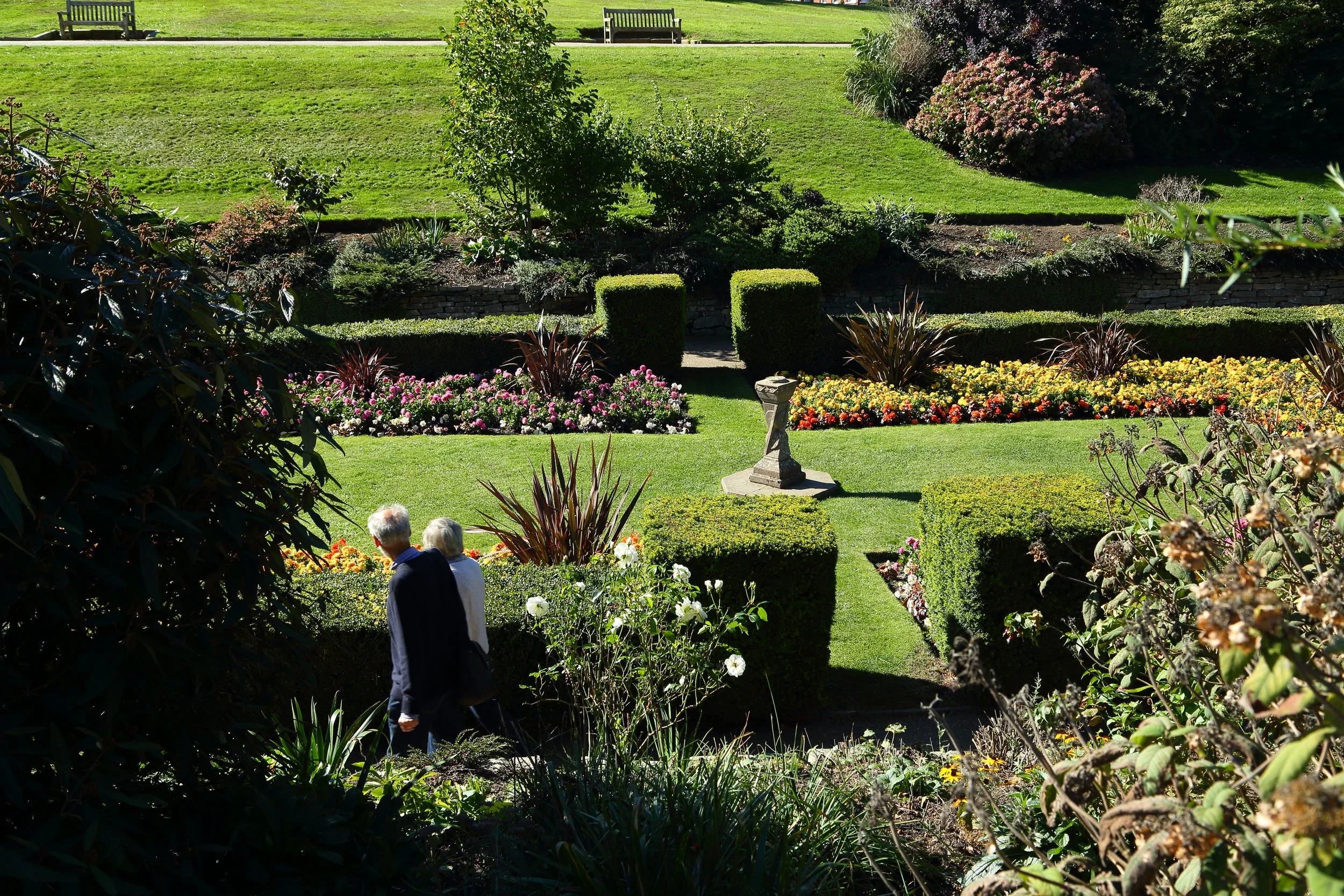 Two elderly people walking through a colorful garden with neatly trimmed bushes, flowering plants, and a stone pedestal in the center.