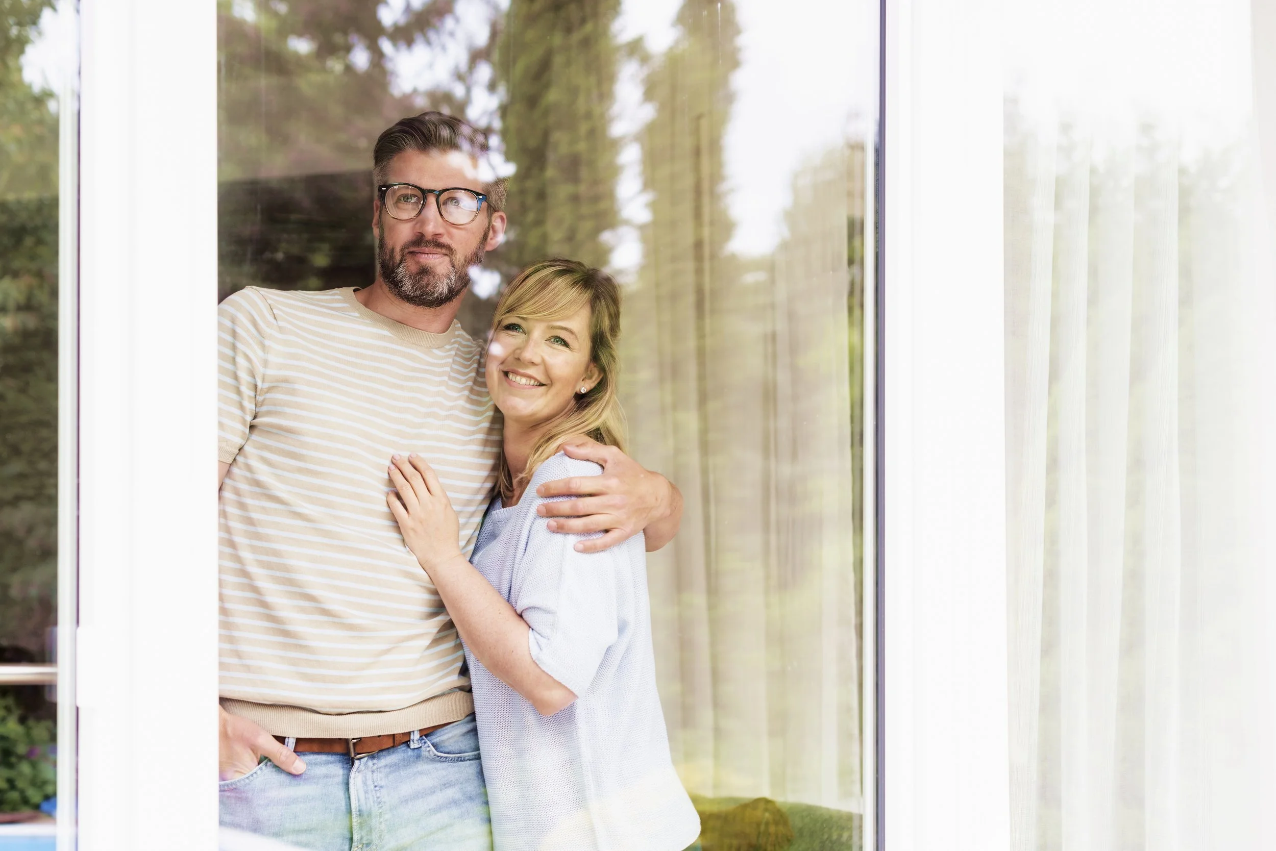 A man with glasses and a beard wearing a striped shirt stands next to a woman in a light-colored sweater, both smiling and embracing each other inside near a large window with curtains, with trees visible outside.