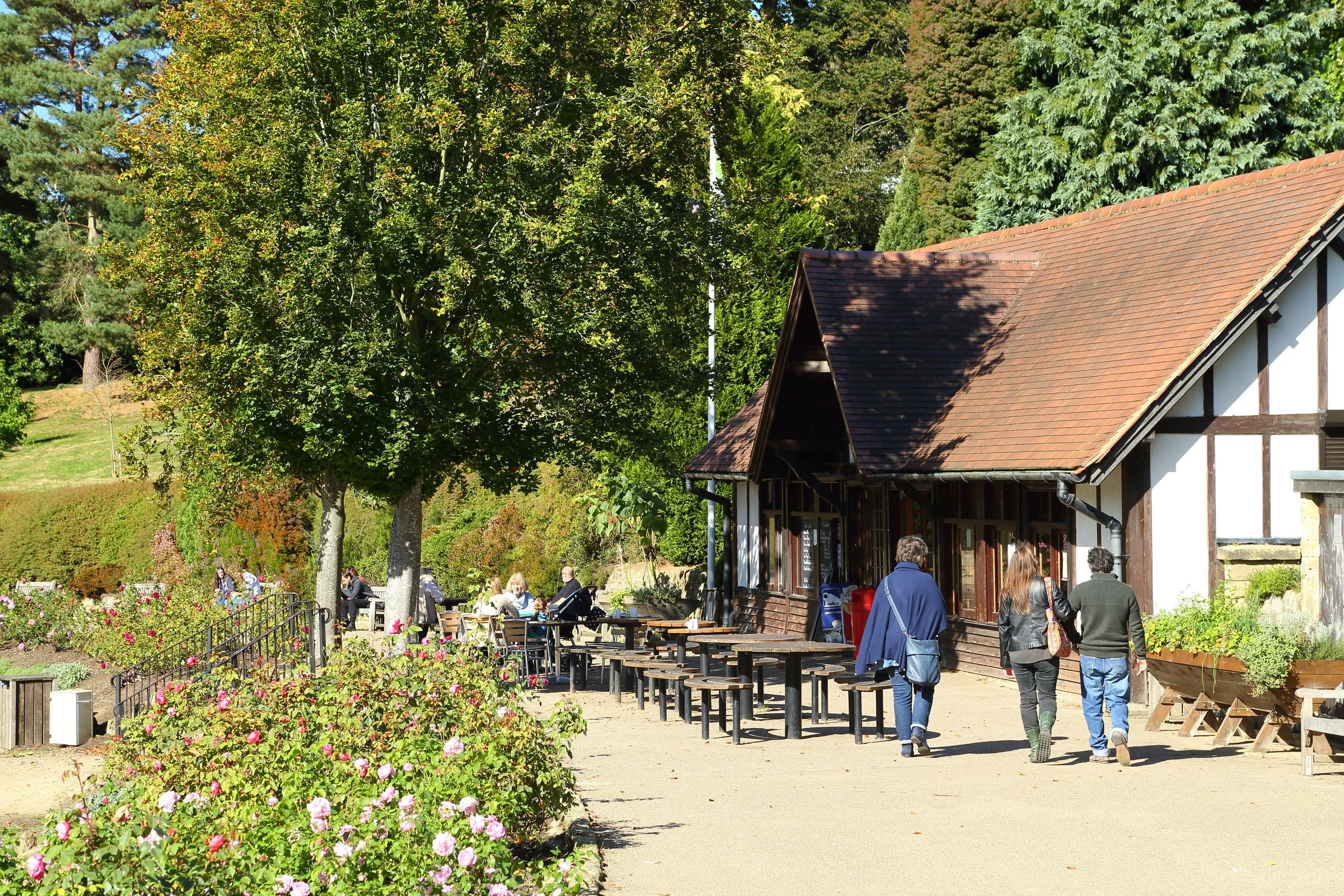 People walking by a rustic building with outdoor seating, surrounded by blooming flowers and green trees on a sunny day.
