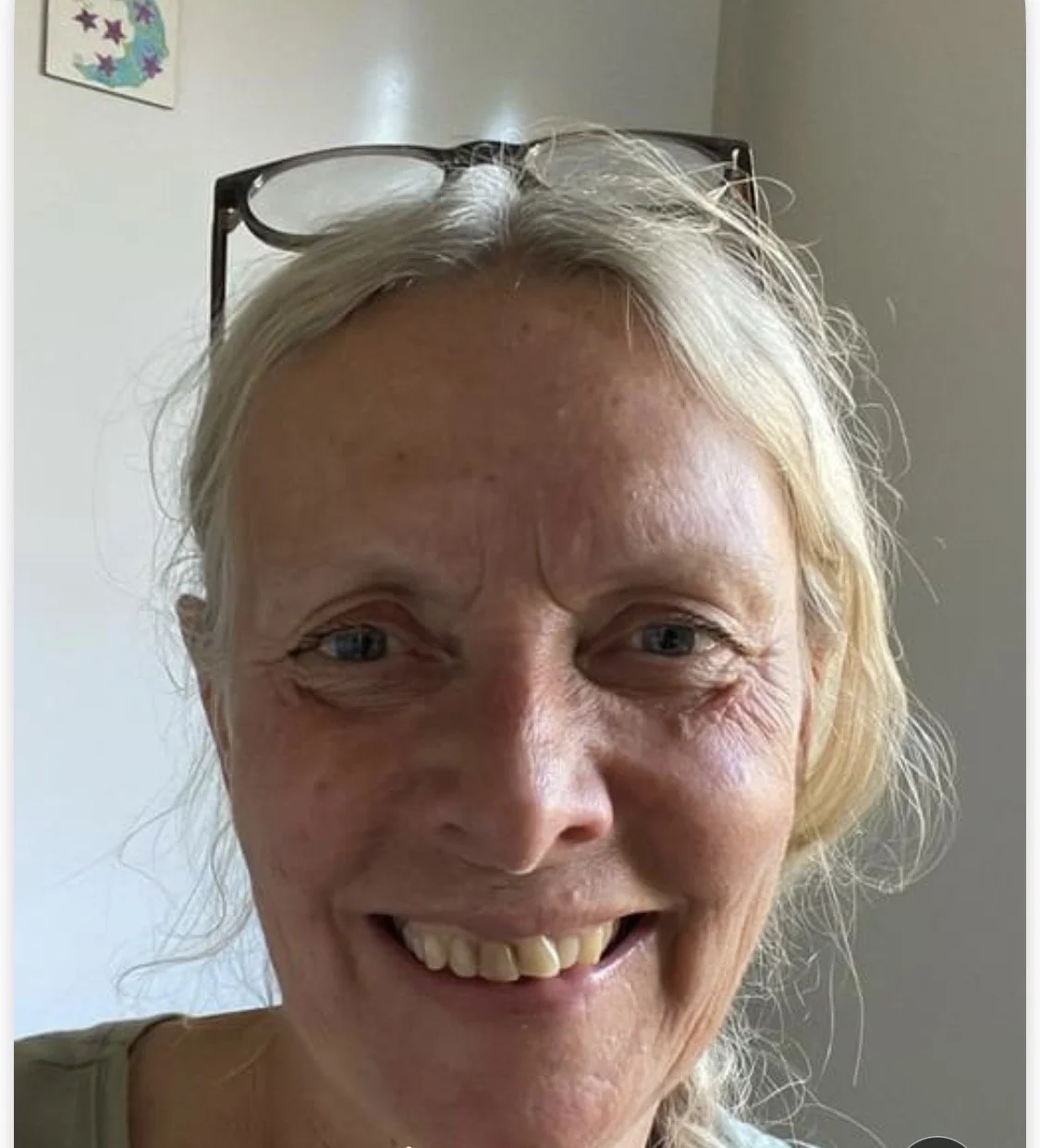 Et smilende ældre kvinde med blondt hår og briller på hovedet, der sidder indendørs.