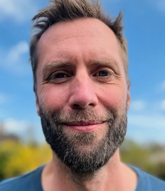 Et close-up portræt af en mand med skæg, gråt hår og smil, baggrund med blå himmel og grønne træer.