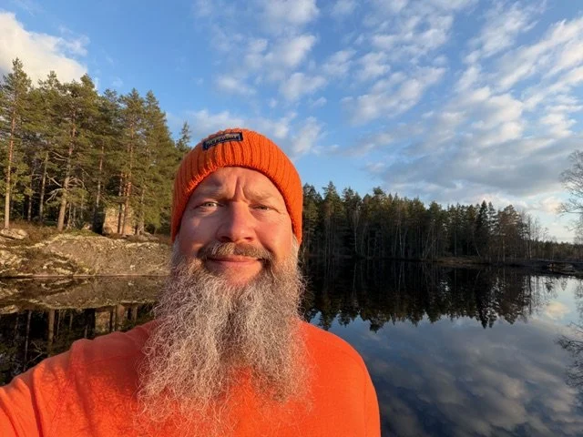En mand med langt gråt skæg iført orange hue og orange jakke står foran en spejlblank sø omgivet af træer under en skyet himmel.