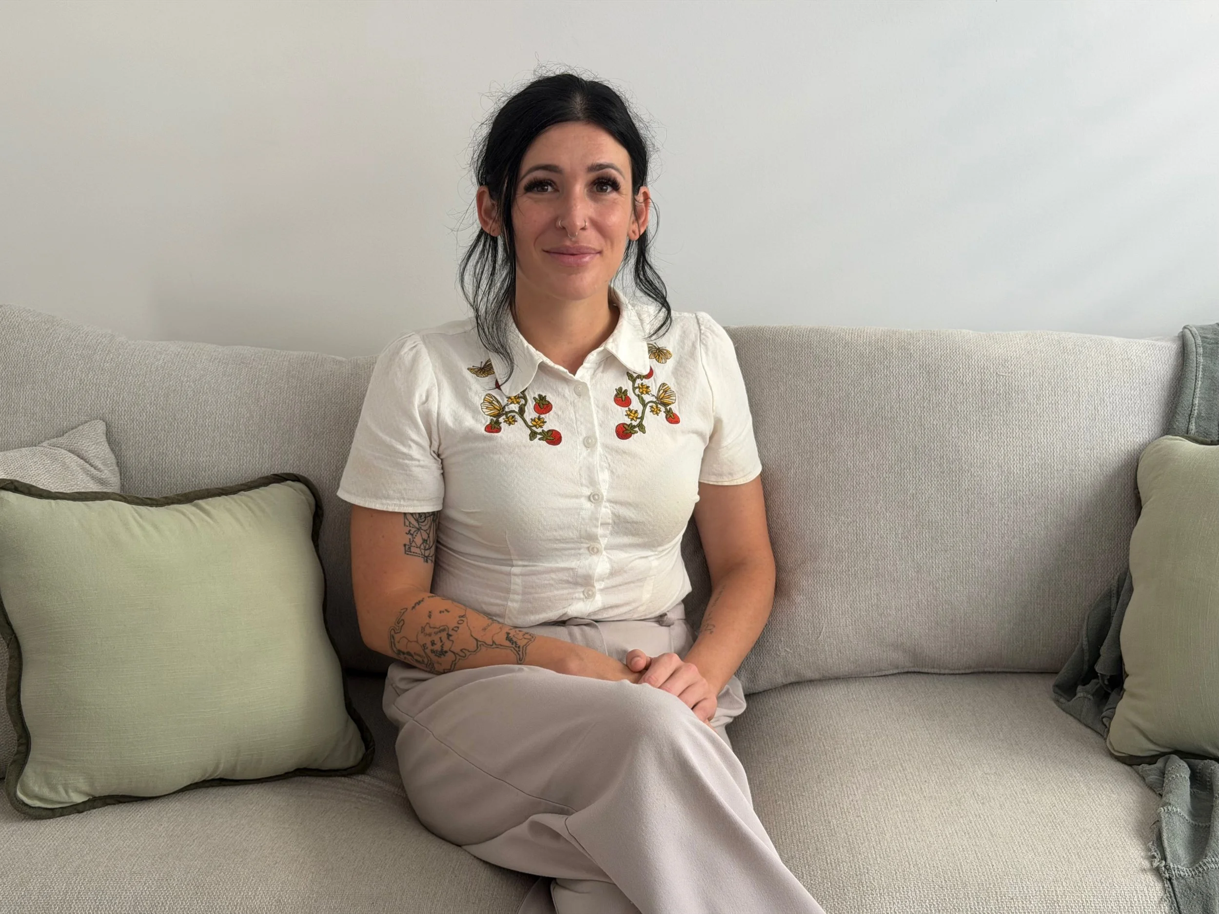 A woman with black hair sitting on a light-colored couch, smiling, wearing a white shirt with embroidered flowers and birds, and beige pants.
