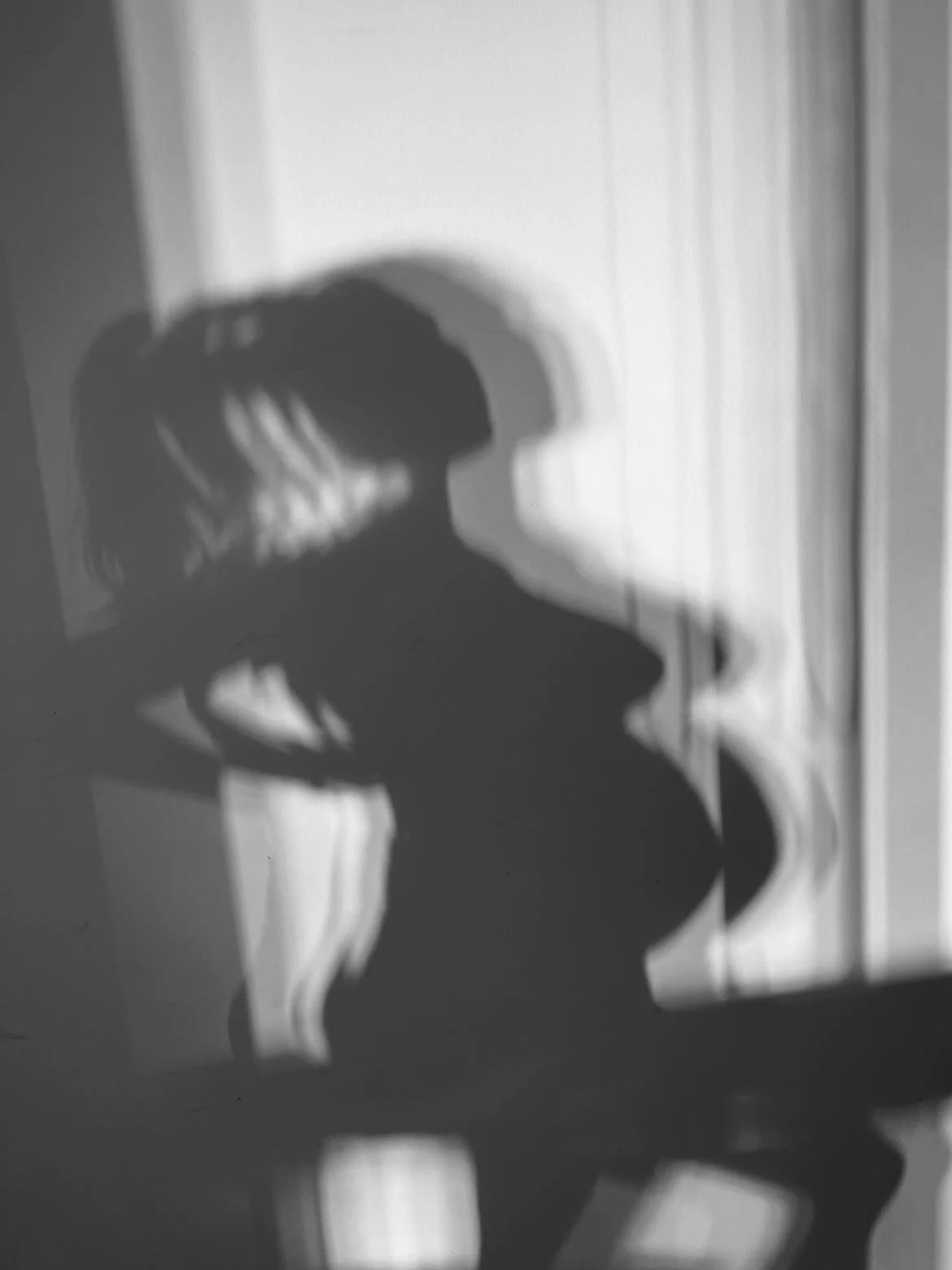 Shadow of a person with long hair and headphones cast on a wall, with vertical blinds partially visible.