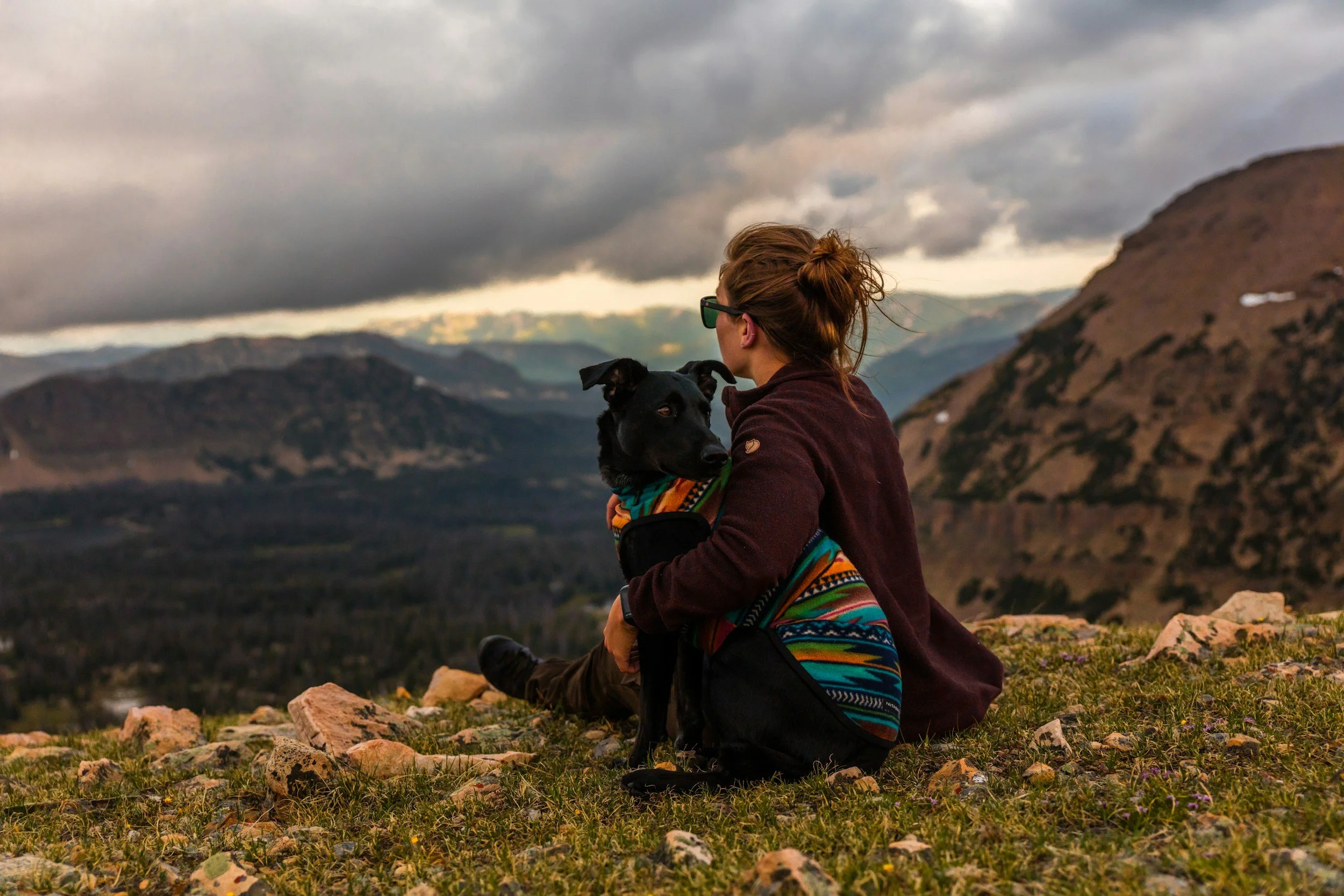 A woman sitting on a grassy mountain ridge holding a black dog, with mountains and cloudy sky in the background.