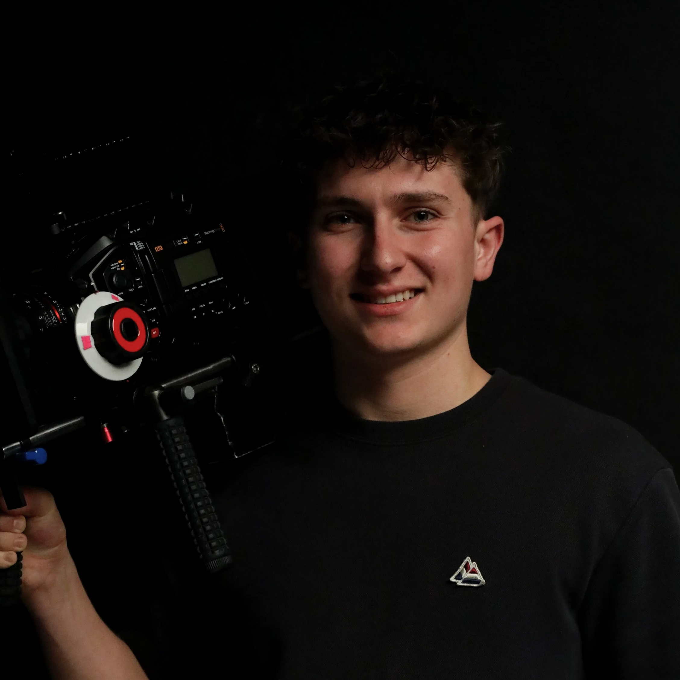 Close-up of a young man with curly hair smiling, holding a professional camera with headphones hanging from it, against a black background.