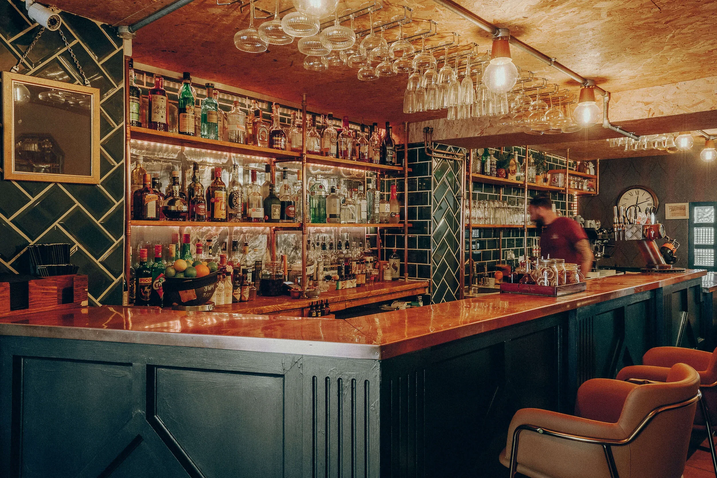 A cozy bar scene with a wooden counter, shelves stocked with various bottles of alcohol, and hanging glassware under warm lighting. There are orange chairs in the foreground and a person behind the bar, slightly blurred.