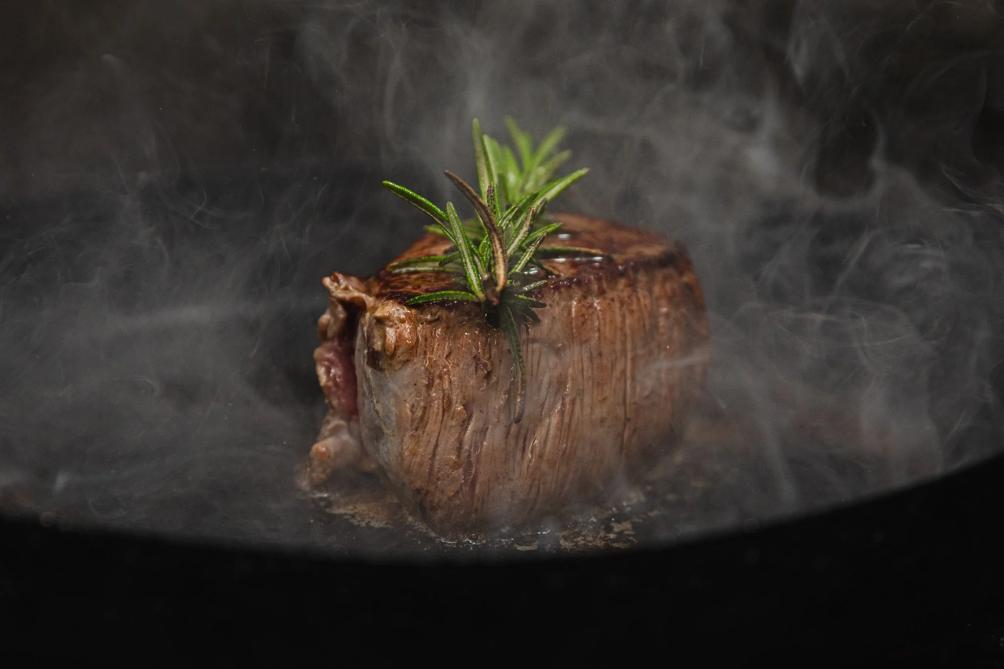 A piece of steak cooking in a pan with steam and fresh rosemary on top.