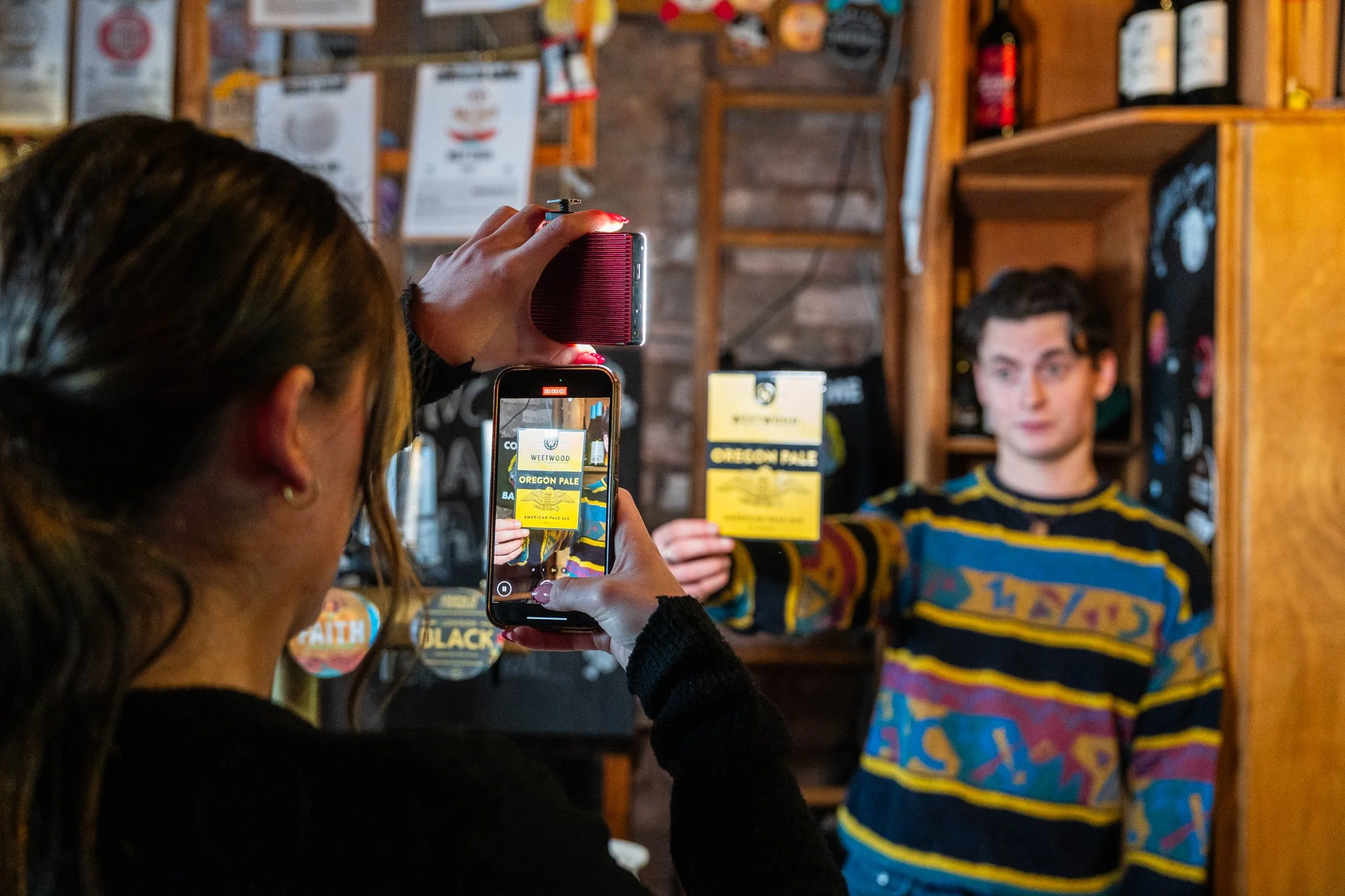 A woman takes a photo of a young man holding a yellow beer label at a craft beer shop. The man wears a colorful patterned sweater, and the woman has long dark hair and is using her phone to capture the scene.