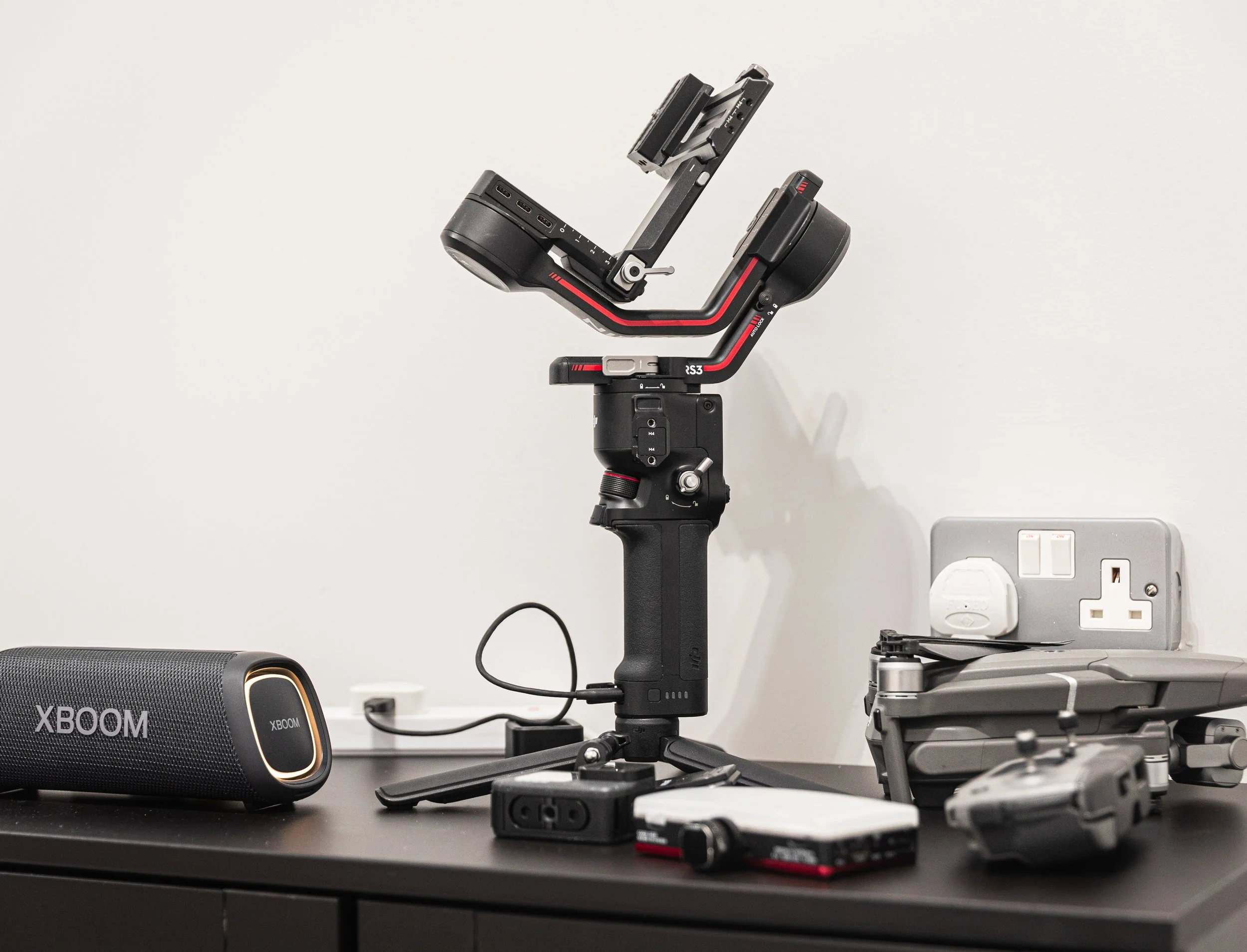 A camera gimbal stabilizer on a black table, with a portable speaker, drone, and electronic accessories nearby, and a white wall with an electrical outlet in the background.