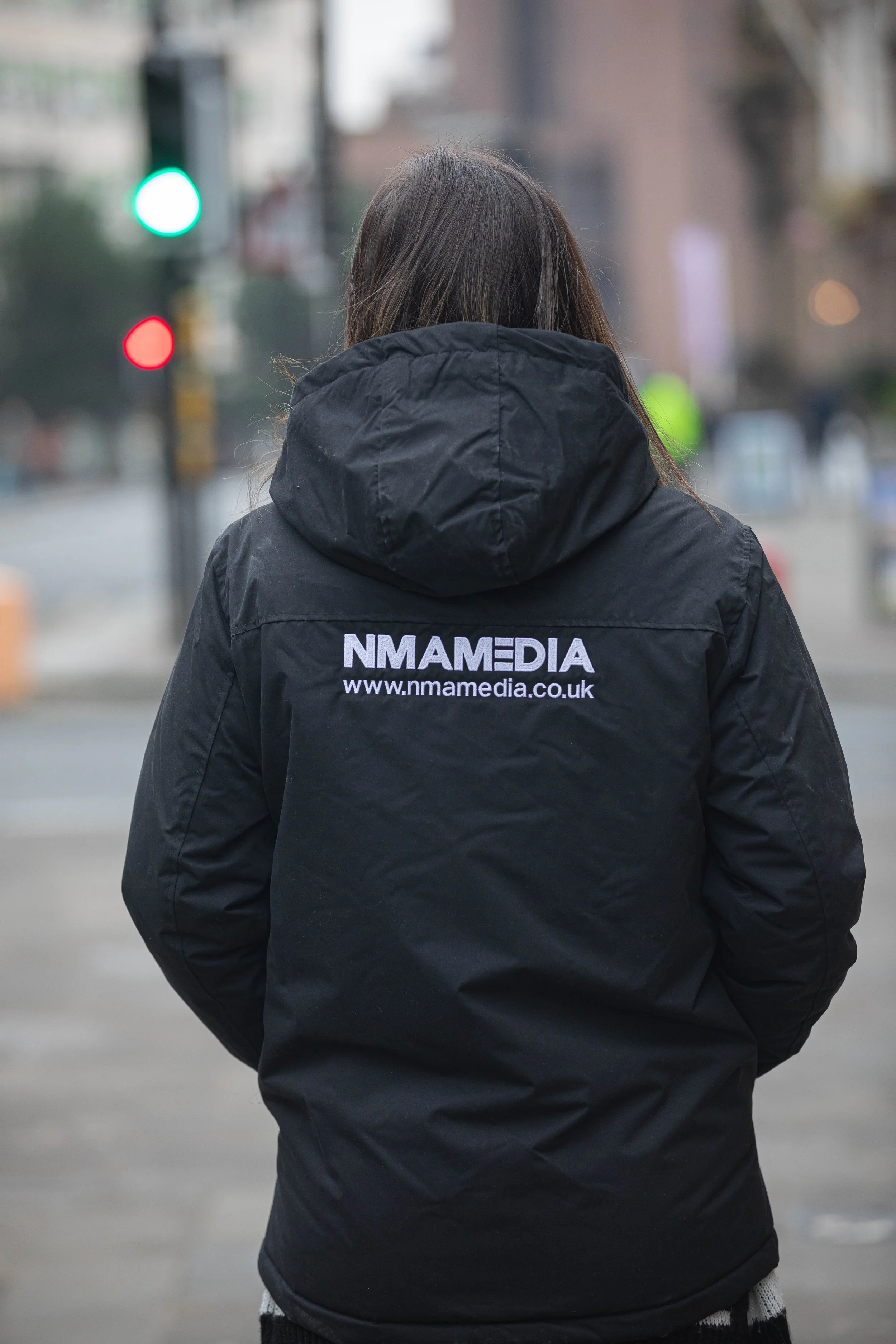 Person wearing a black hoodie with 'NMAMEDIA' and website on the back, walking outdoors in an urban area.