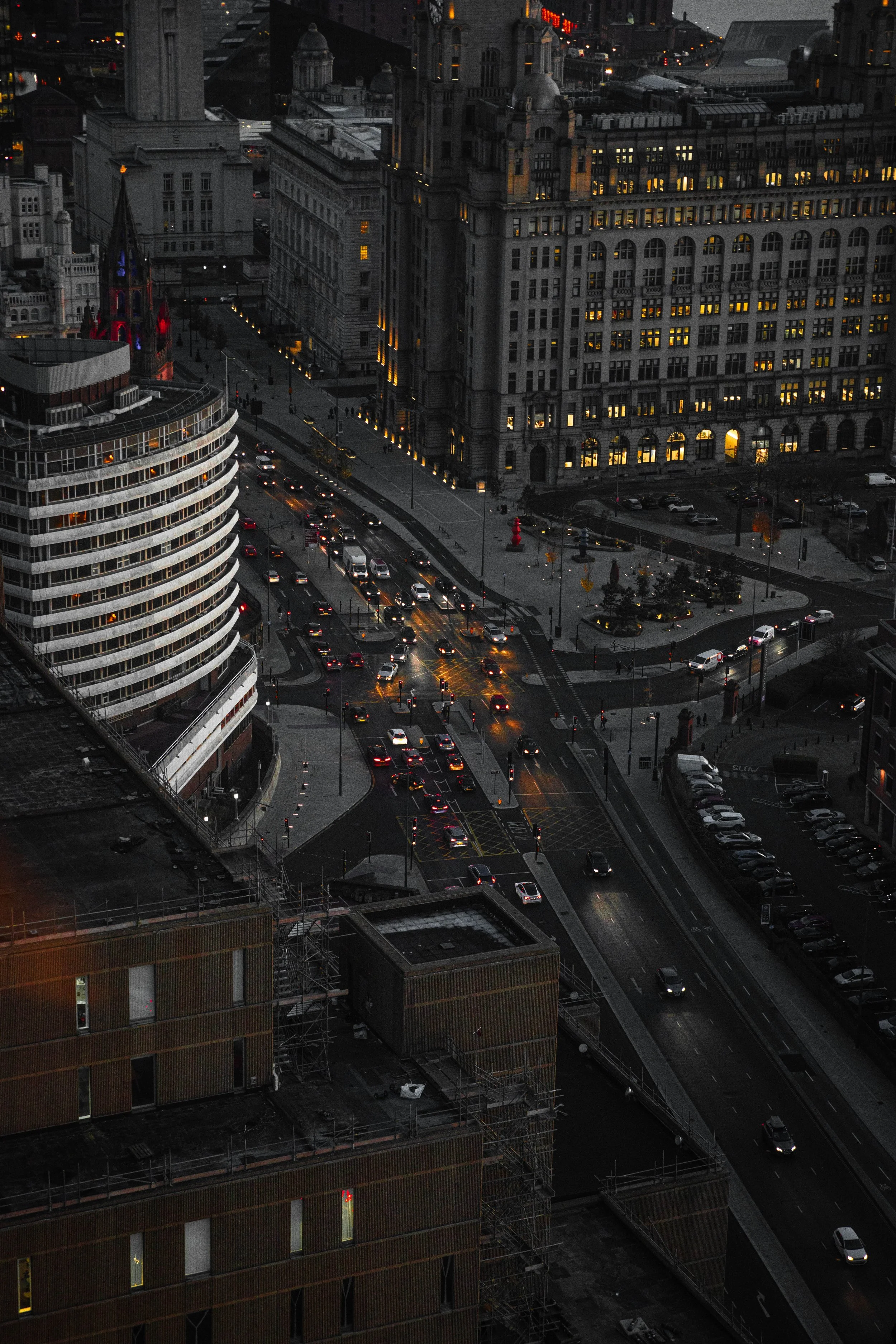 Nighttime cityscape showing busy streets with cars, illuminated buildings, and a park with trees and a red sculpture.