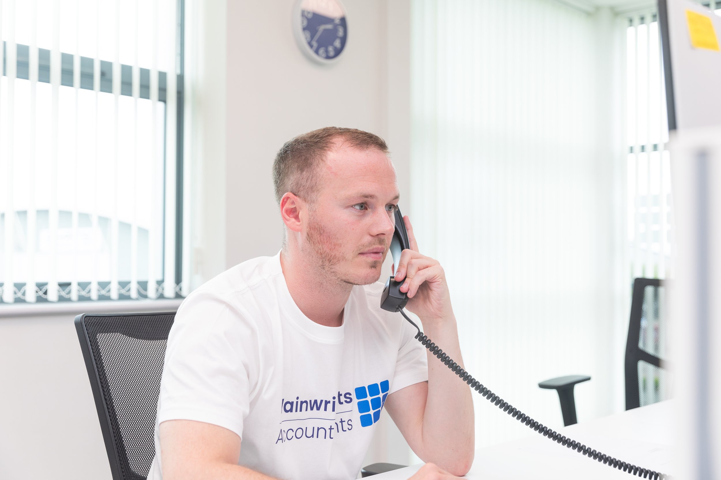 A man with short hair and a white t-shirt talking on a corded telephone in an office setting.