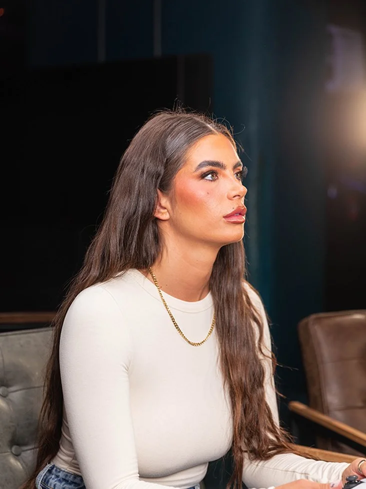 A young woman with long brown hair, wearing a white long-sleeve top, gold chain necklace, and makeup, is sitting at a table in a room with dark walls and a light source to her right.